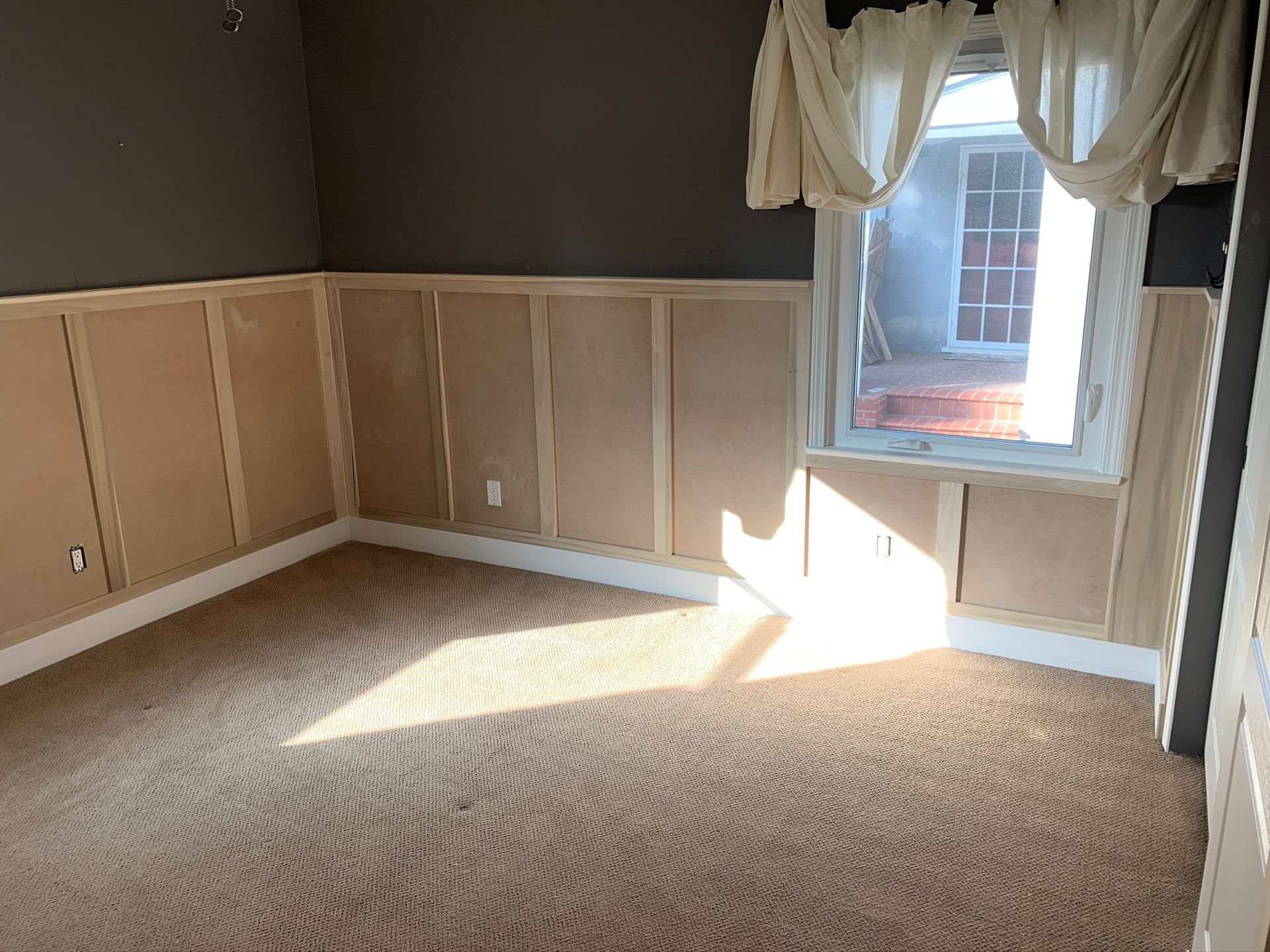 Empty room with carpet, paneled walls, and window with curtains; sunlight streams in.
