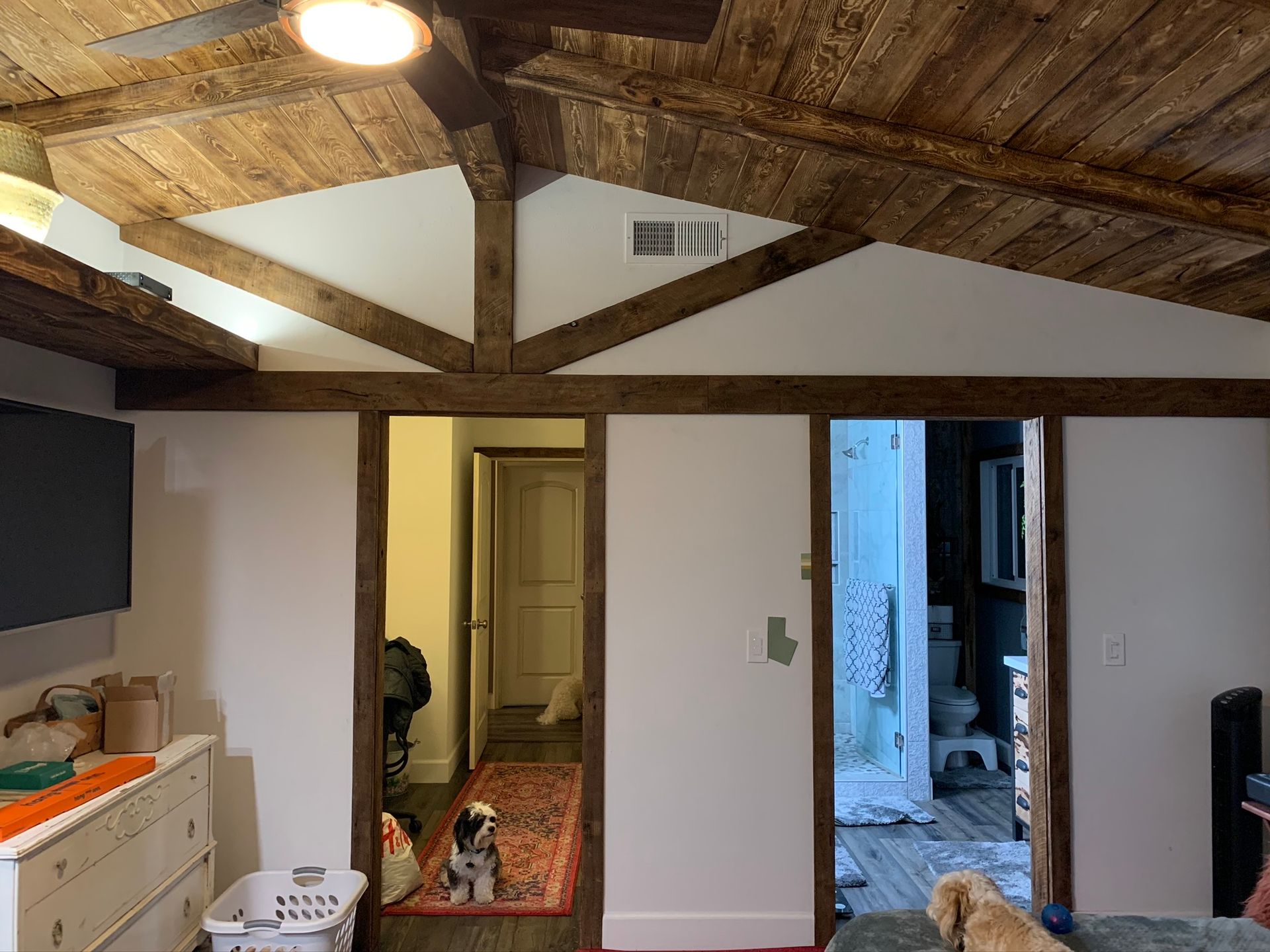 Wooden-beamed ceiling and trim frame doorways and an open area with a dresser and laundry basket. A dog sits on a rug.