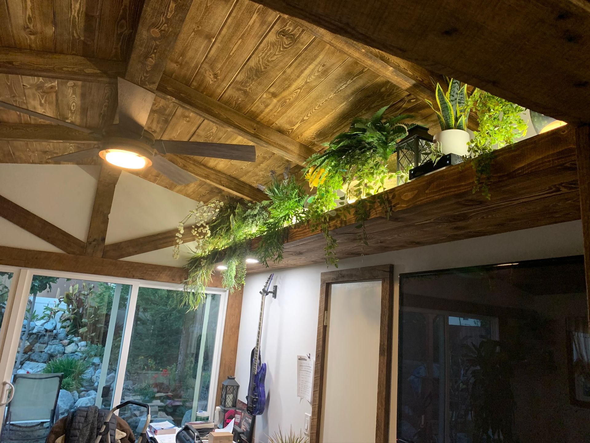 Wooden ceiling with plants and a ceiling fan. Bright lighting illuminates the greenery and rafters.