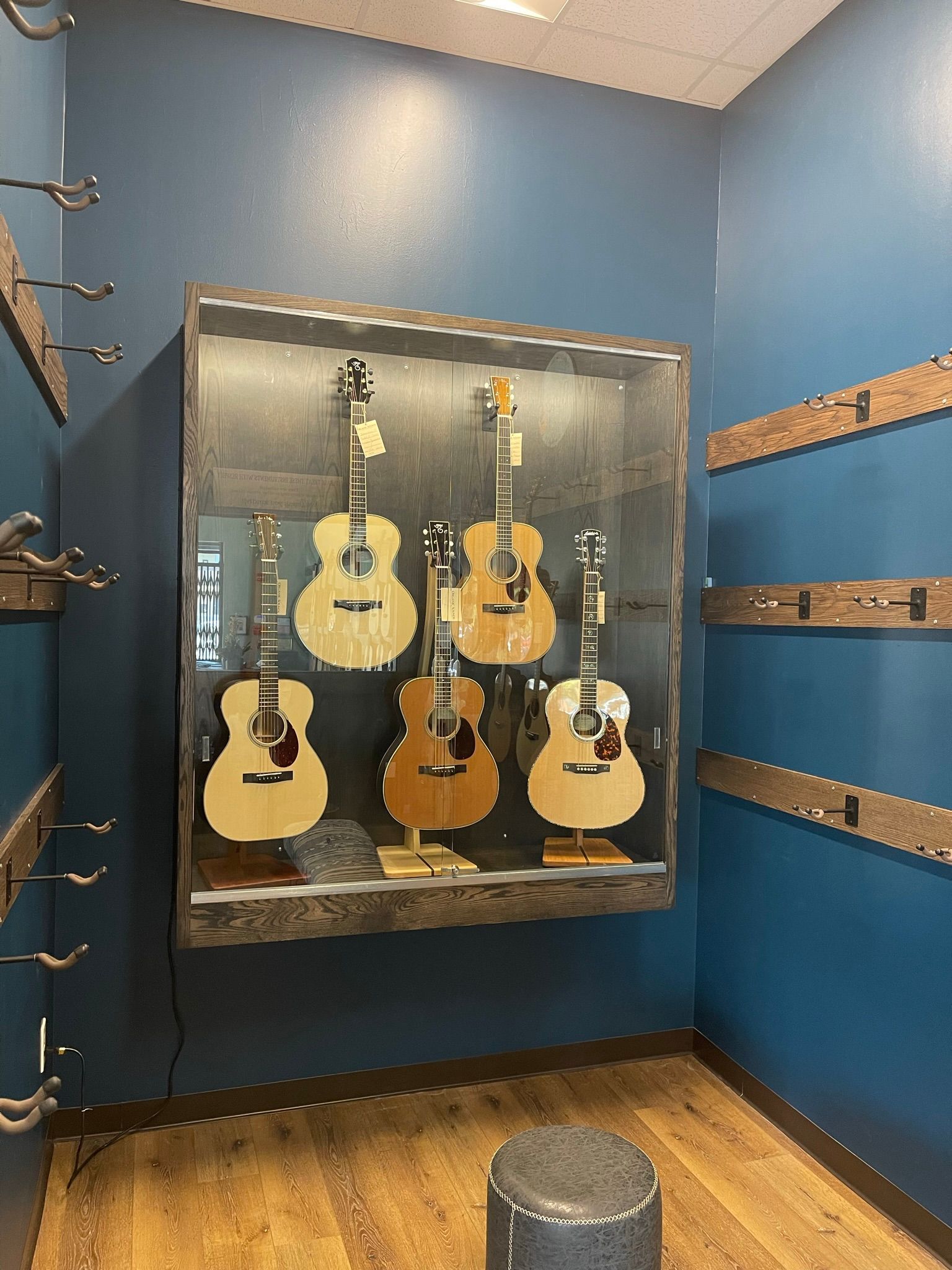 Guitars displayed in a glass case on a blue wall in a music store. Wooden floor and guitar hangers.