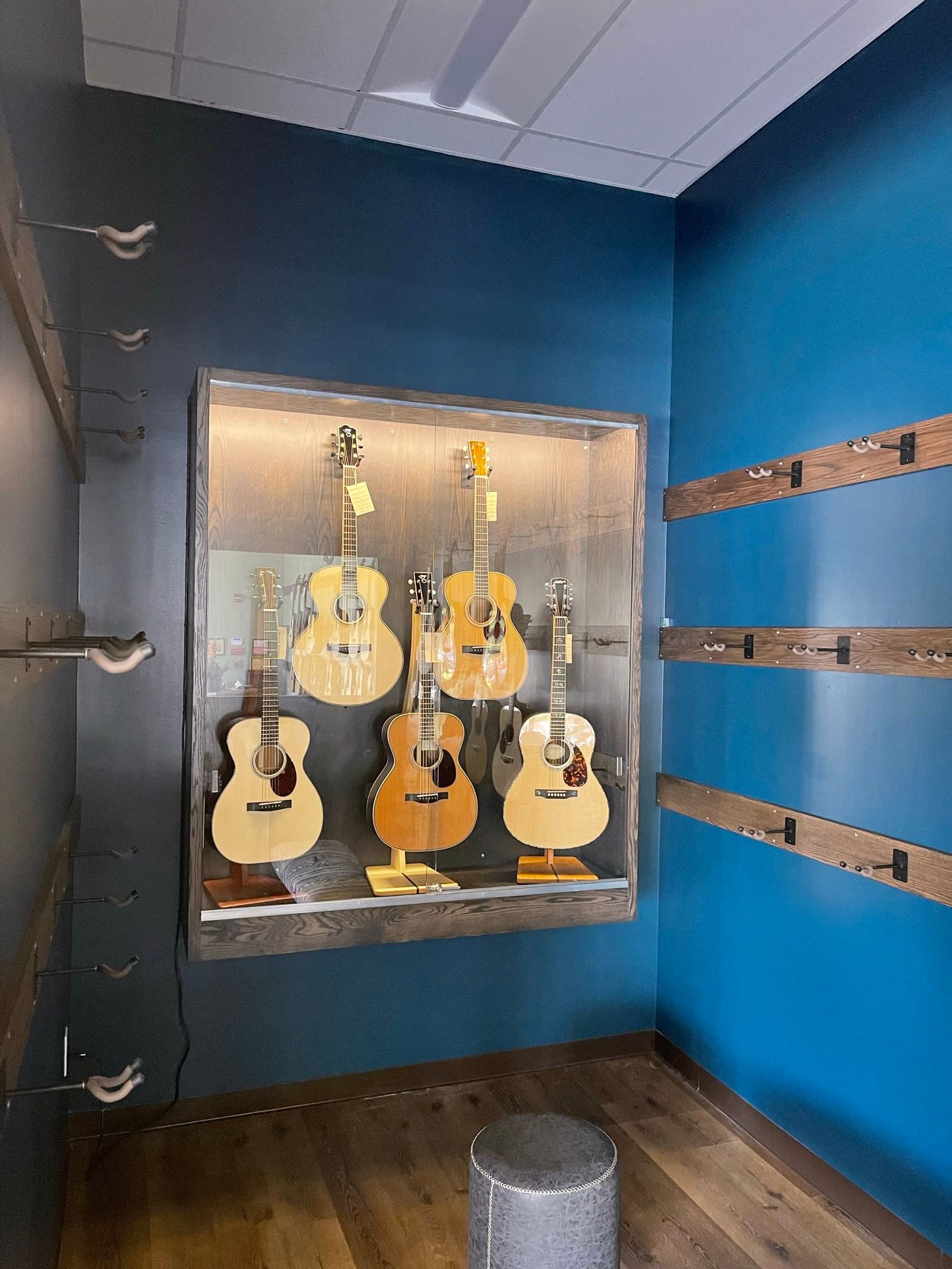 Guitars on display in a glass case on a blue wall, with wooden accents and a stool.