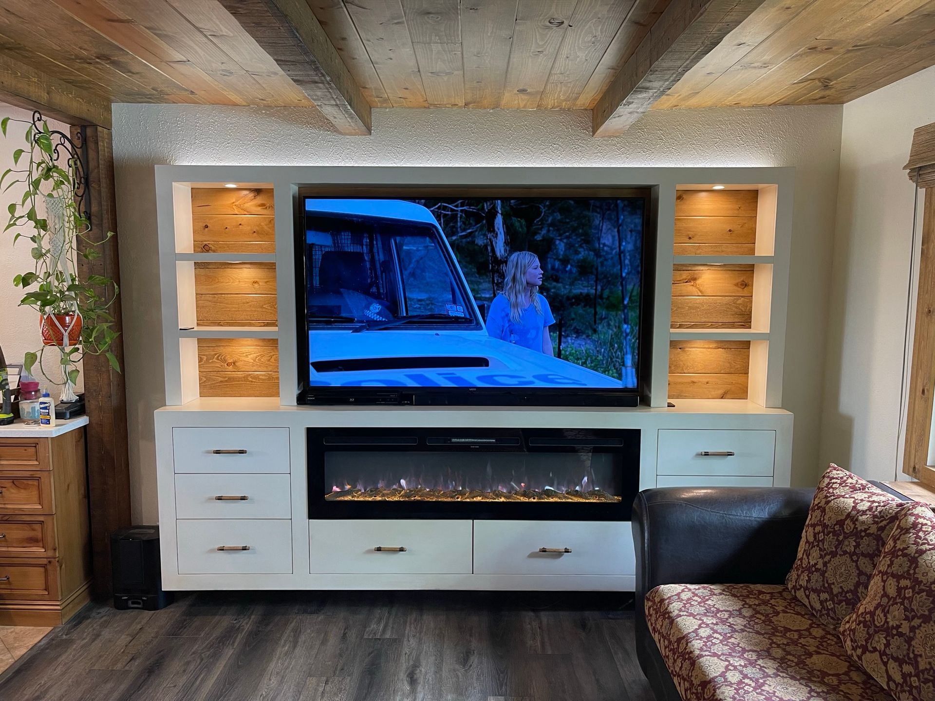 Living room with built-in entertainment center; TV, electric fireplace, shelves, drawers, wood beams, and couch.