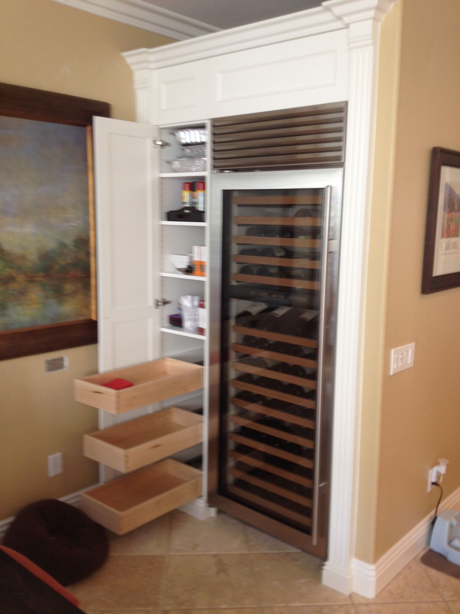 Built-in wine refrigerator and pantry cabinet in a kitchen. White cabinetry, stainless steel fridge, wooden drawers.