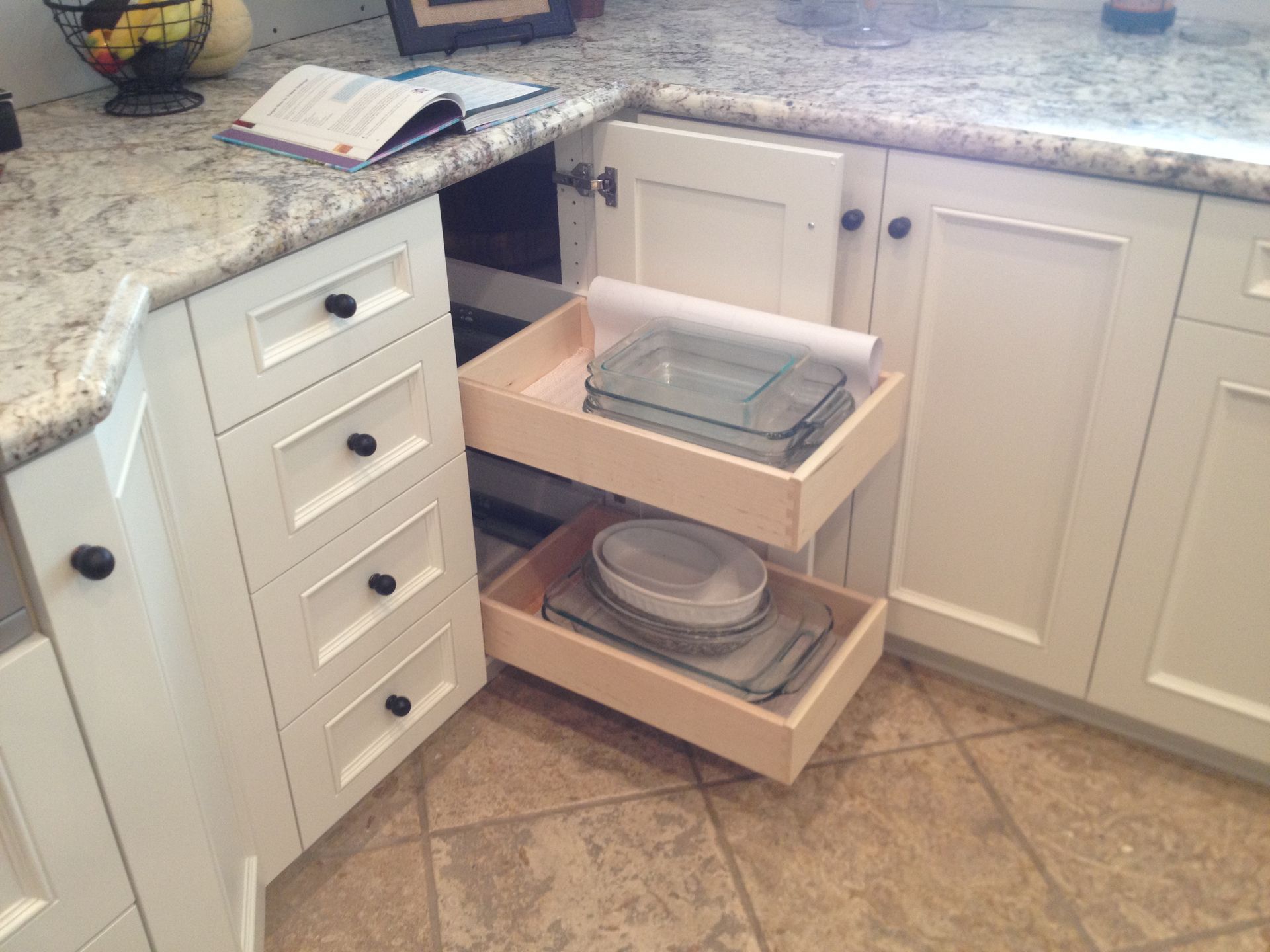 Corner kitchen cabinet with pull-out drawers, containing storage dishes, next to other white cabinets and granite countertop.