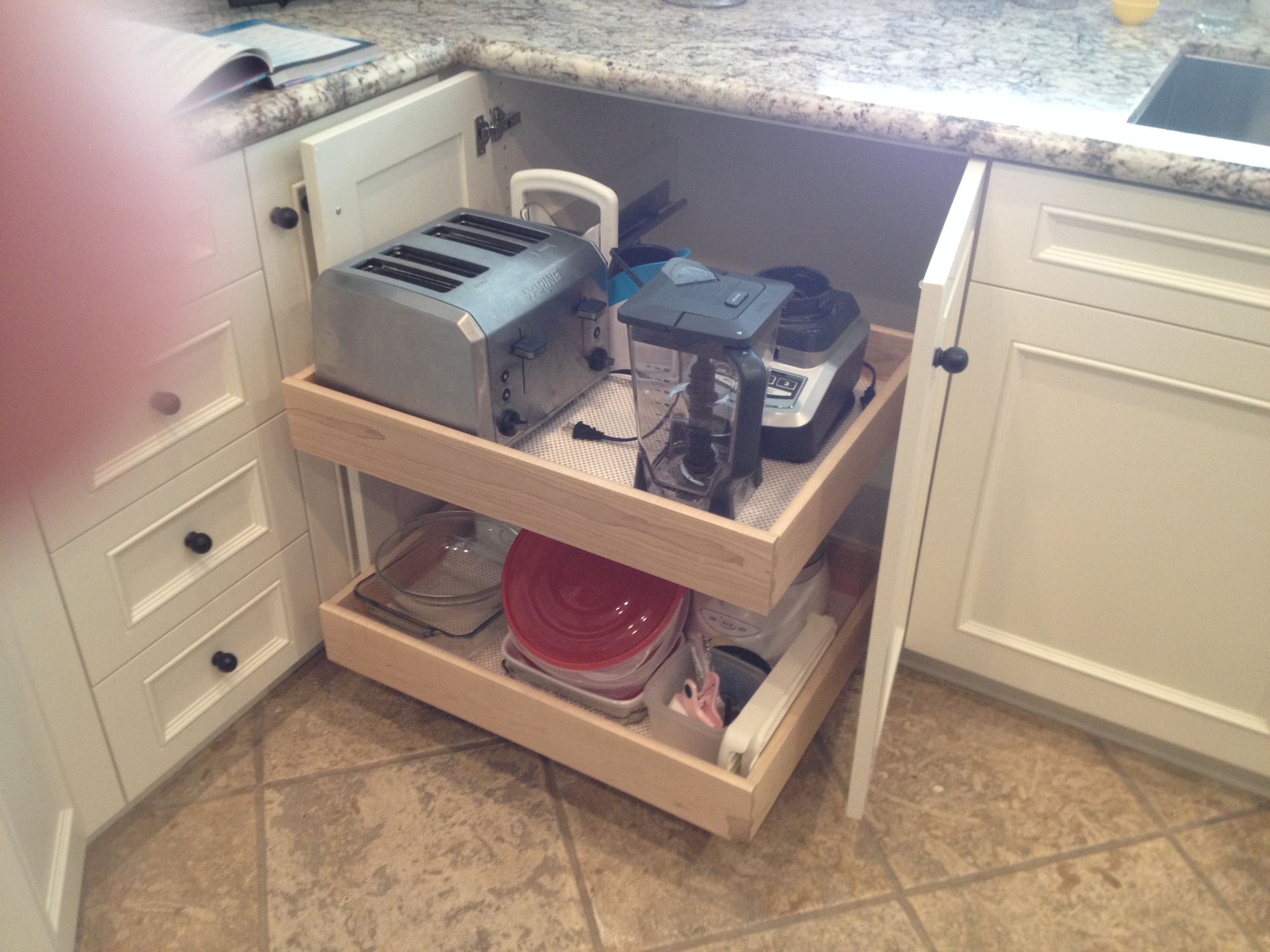 Pull-out shelves inside a white kitchen cabinet, holding a toaster, blender, and containers.