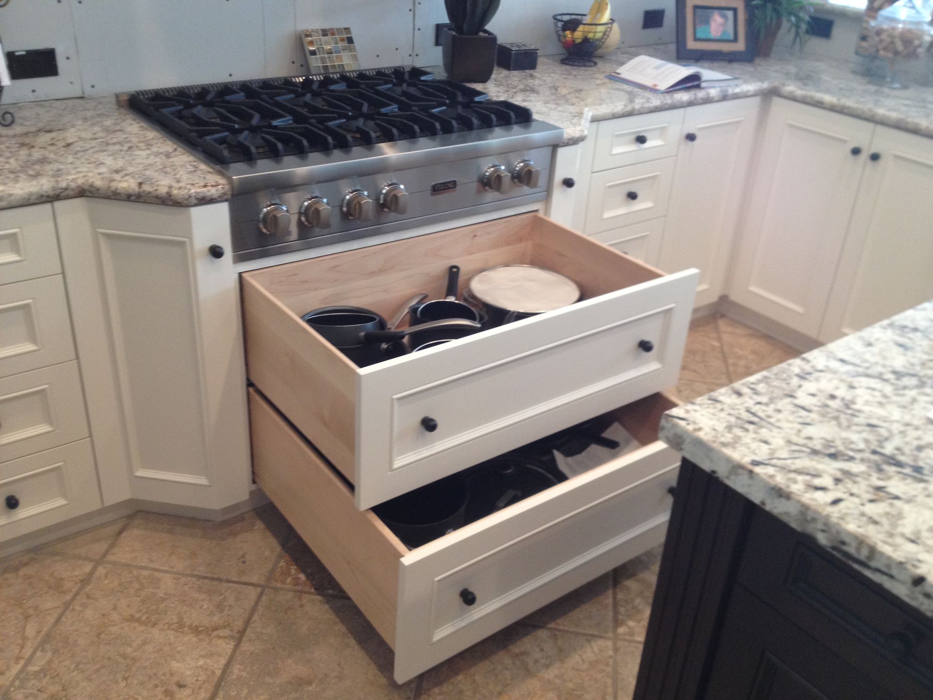 Kitchen with open drawers beneath a stovetop, containing cookware. White cabinets, granite countertop, tile floor.