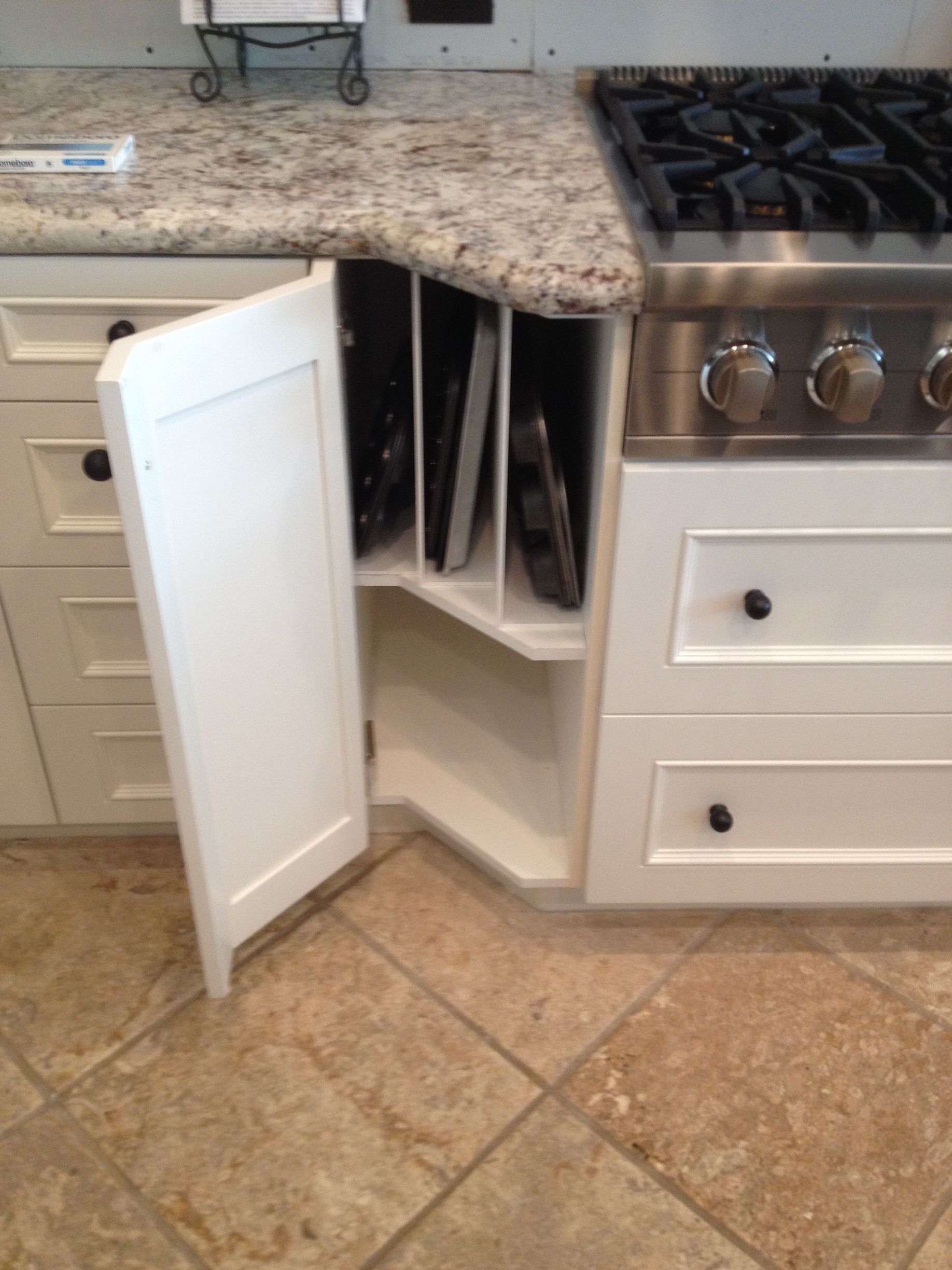 White kitchen cabinet with open door revealing vertical dividers for storing baking sheets next to a stovetop.