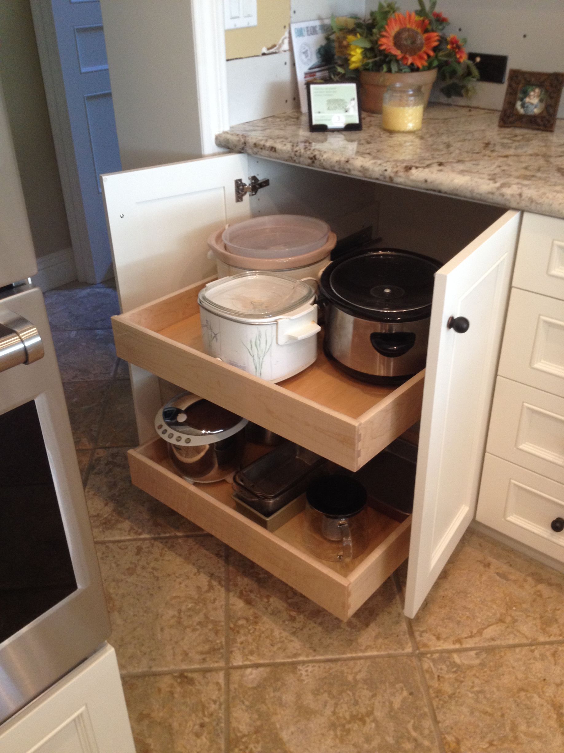 Pull-out kitchen cabinet with two shelves holding slow cookers and containers, open next to a stove.