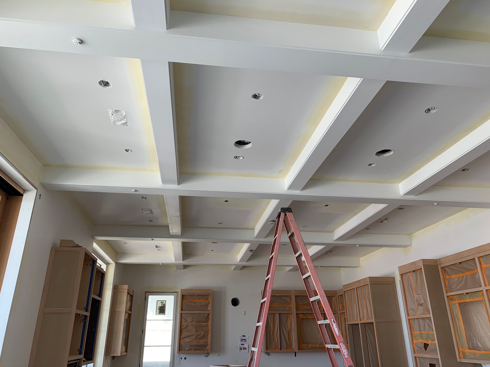 Kitchen ceiling with white beams, recessed lighting, and unfinished cabinetry. Ladder is in the center.