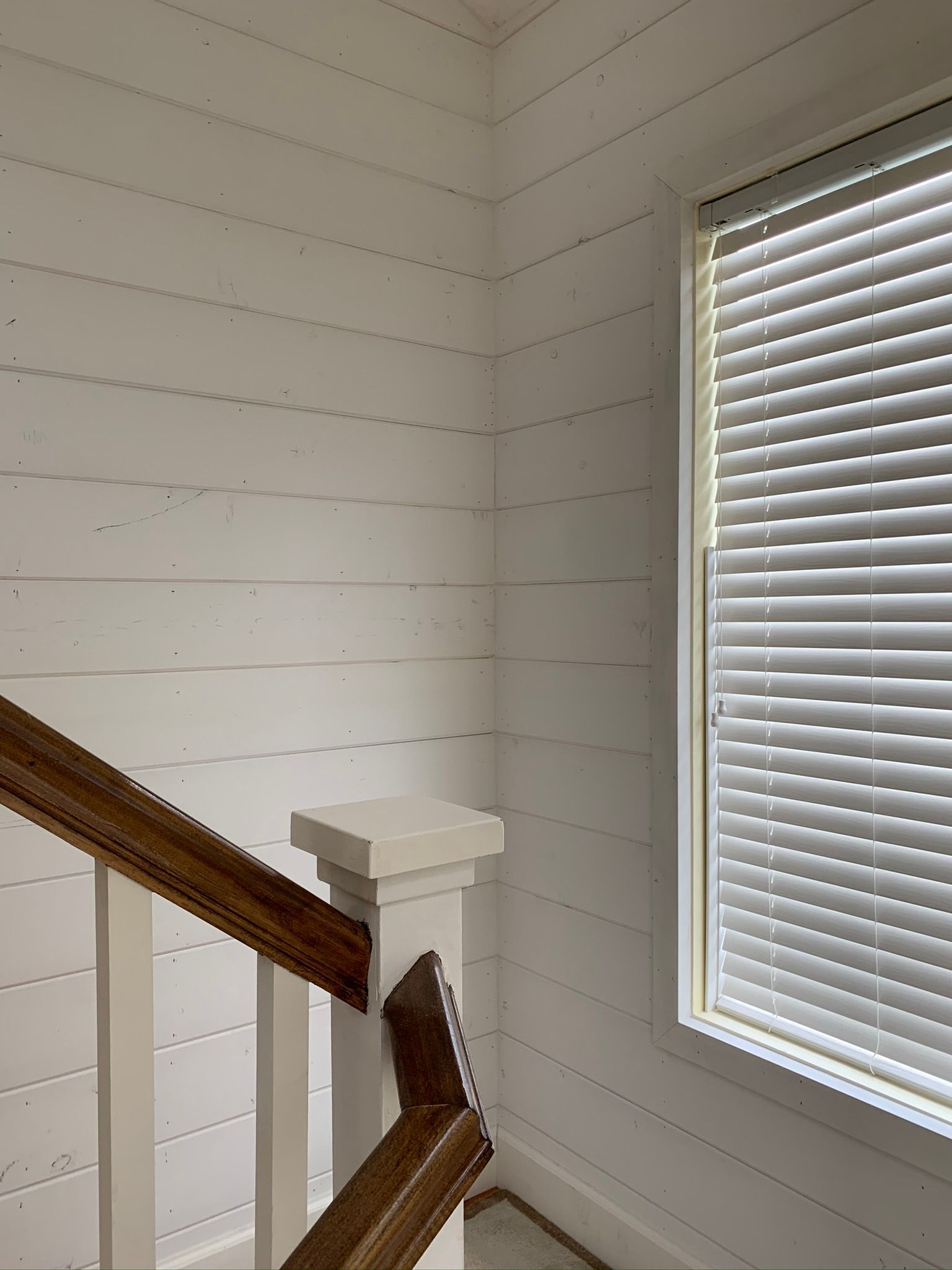 White planked walls next to a window with blinds; wooden banister in the corner.