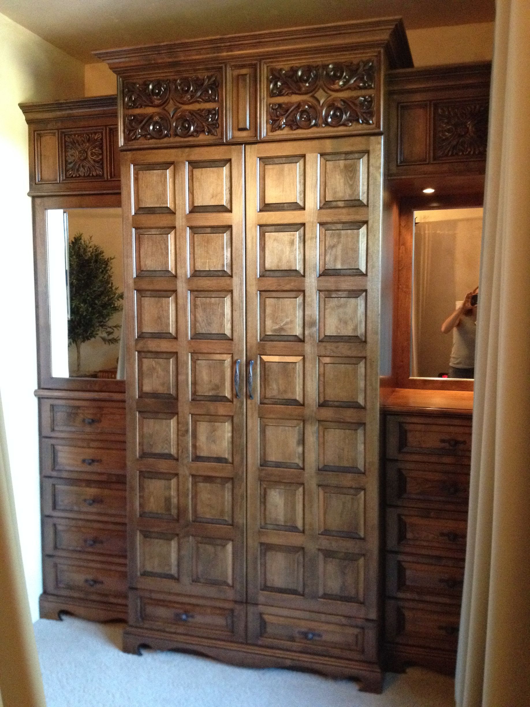 Wooden armoire with carved details, flanked by dressers and mirrors in a room.