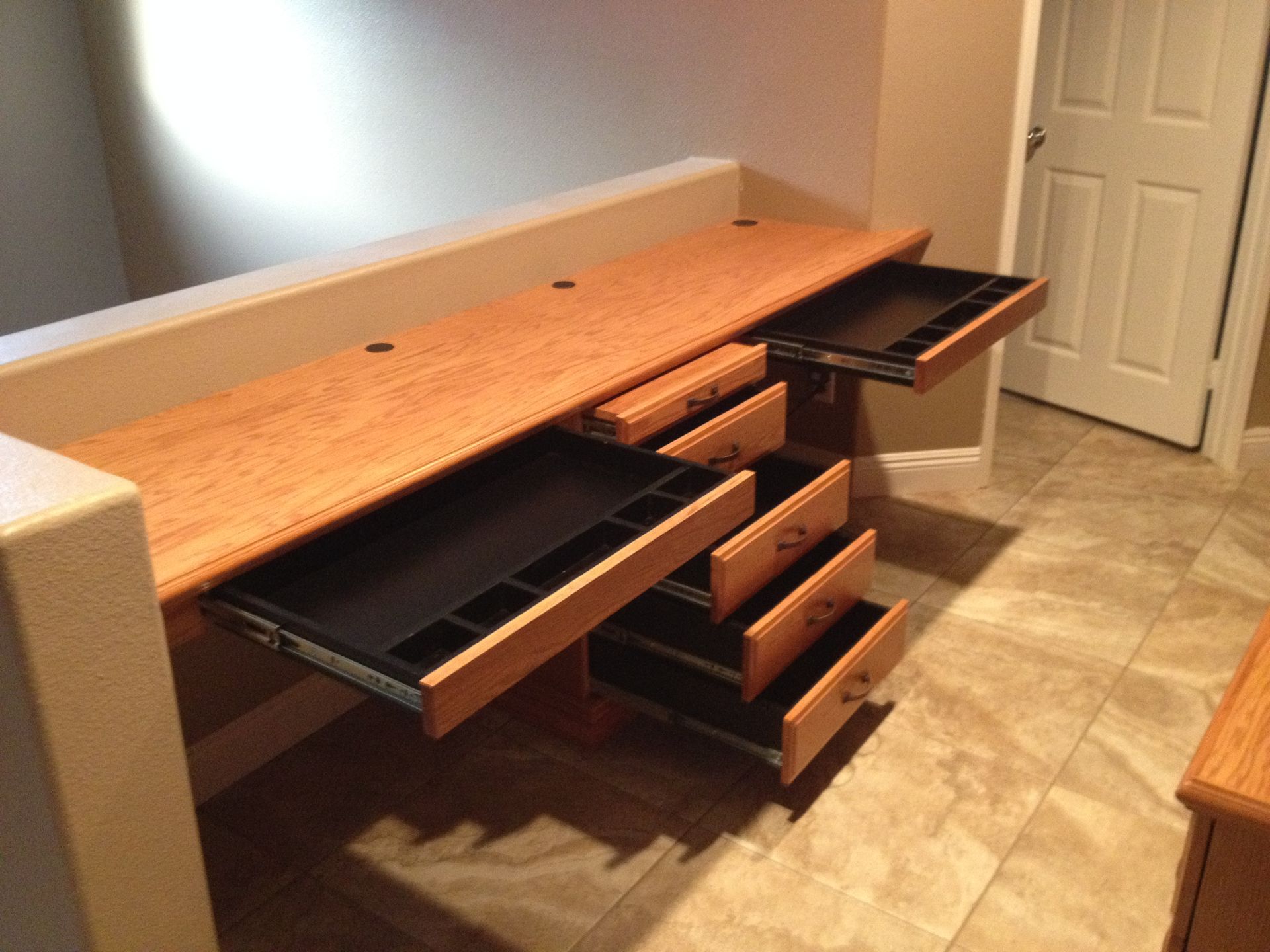 Built-in wooden desk with several open drawers of varying sizes against a beige wall and tile floor.