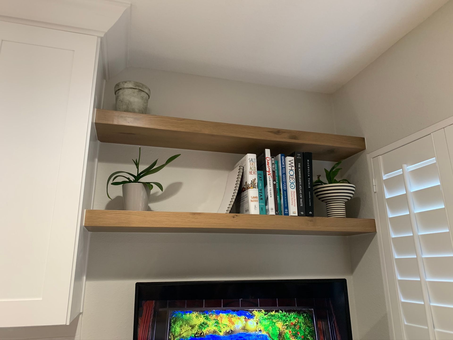 Two wooden shelves with books and plants, mounted above a TV. White cabinetry and shutters are nearby.