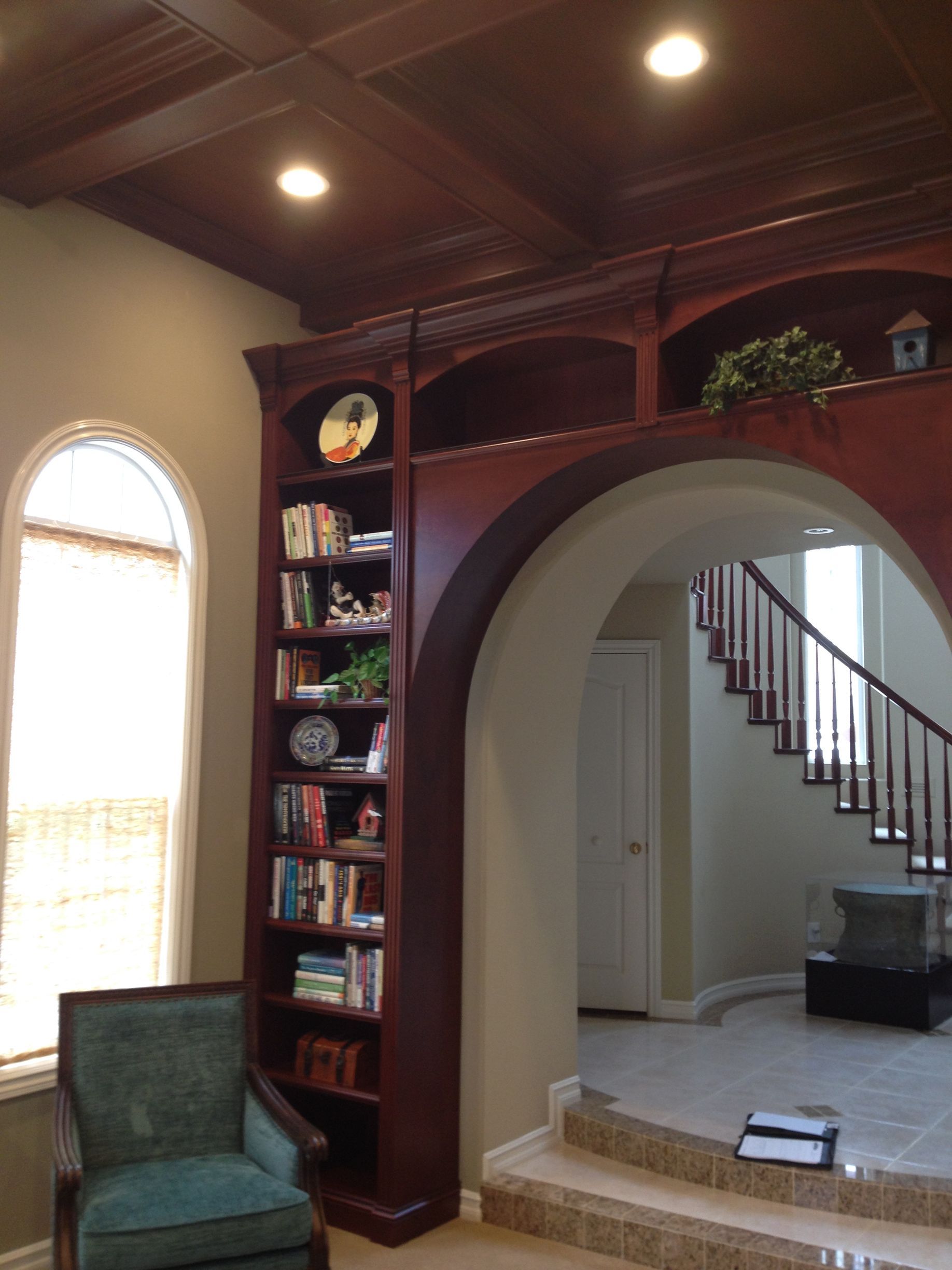 Built-in mahogany bookcase with arched doorway, leading to a staircase. Teal chair by an arched window.