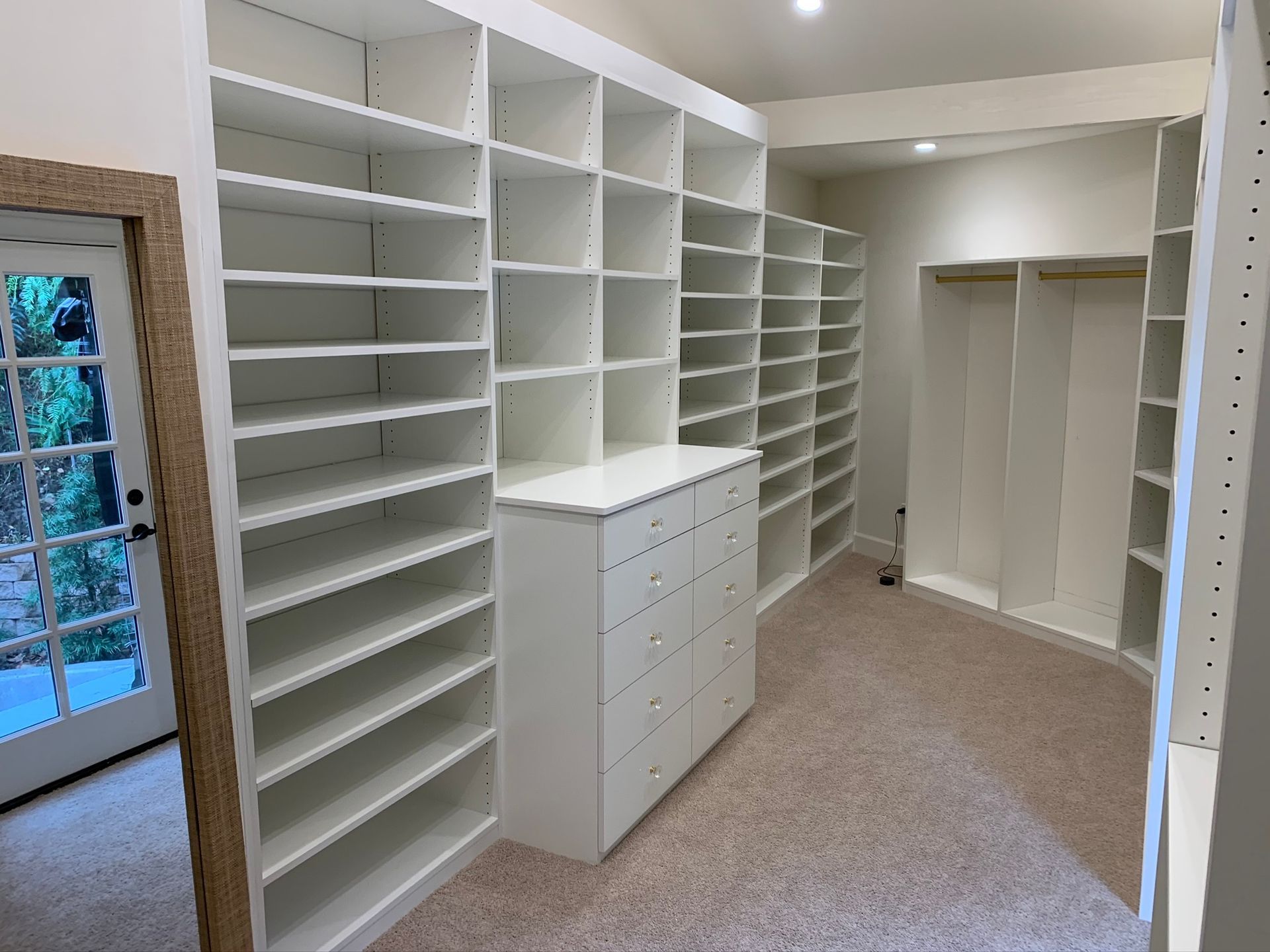 Walk-in closet with white shelving, a chest of drawers, and a beige carpet. A mirror and glass door are on the left.
