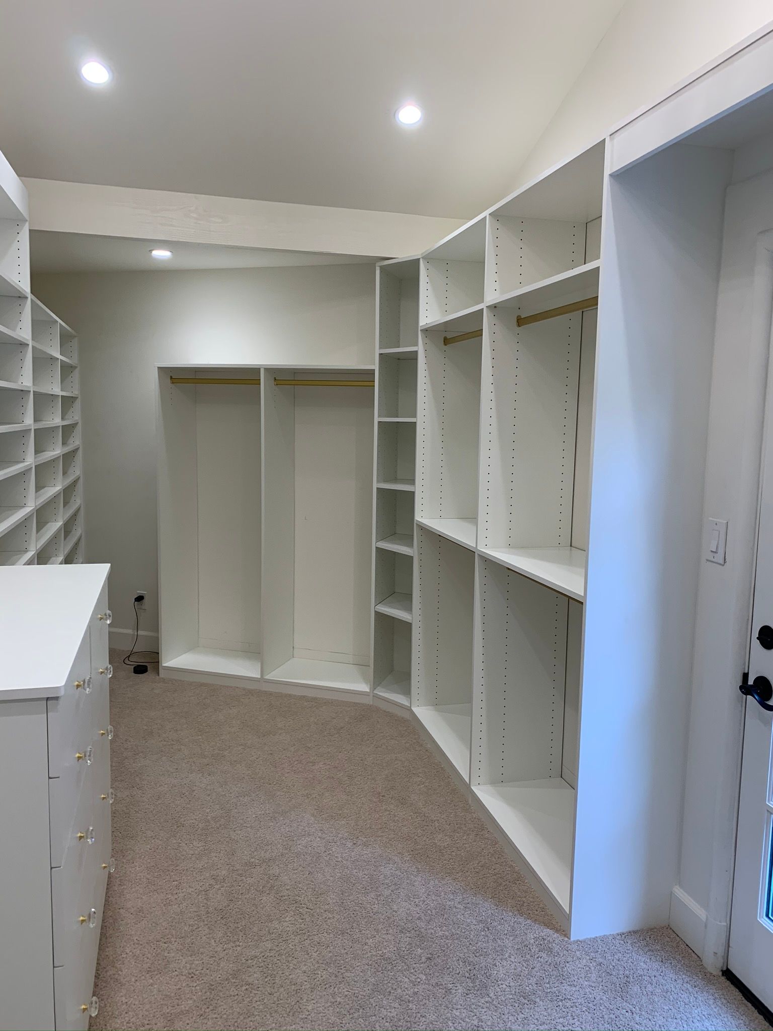 Empty white walk-in closet with shelves and rods, beige carpet, and a door on the right.