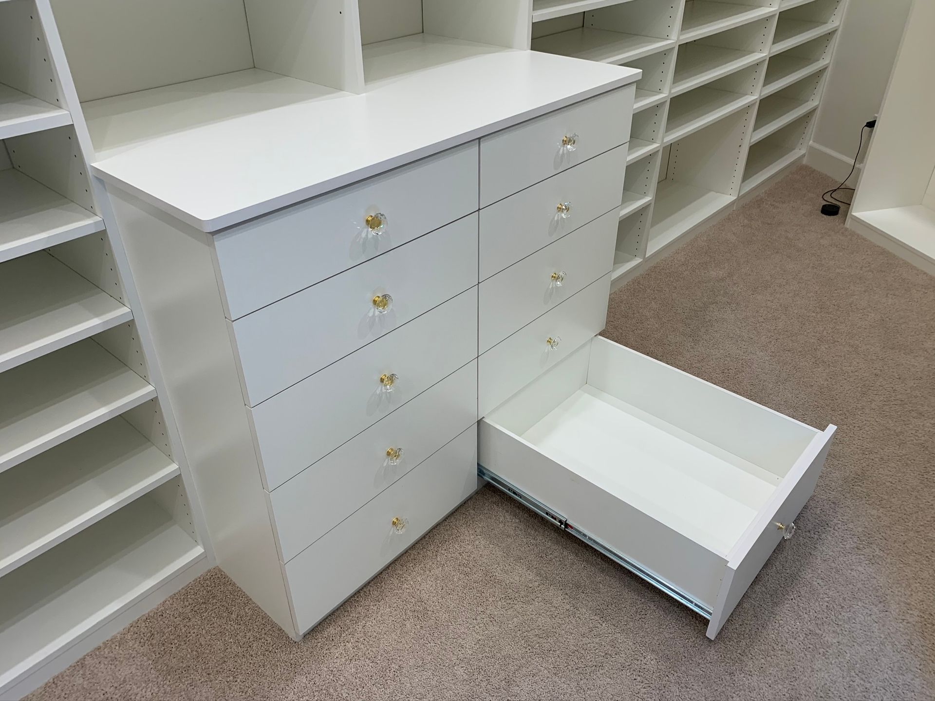 White cabinet with eight drawers, one open, in a closet with shelves; gold knobs.