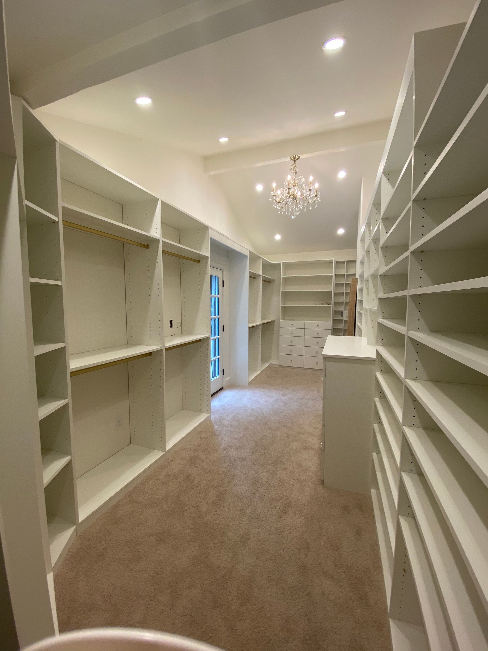 Spacious walk-in closet with white shelves, a chandelier, and beige carpet.