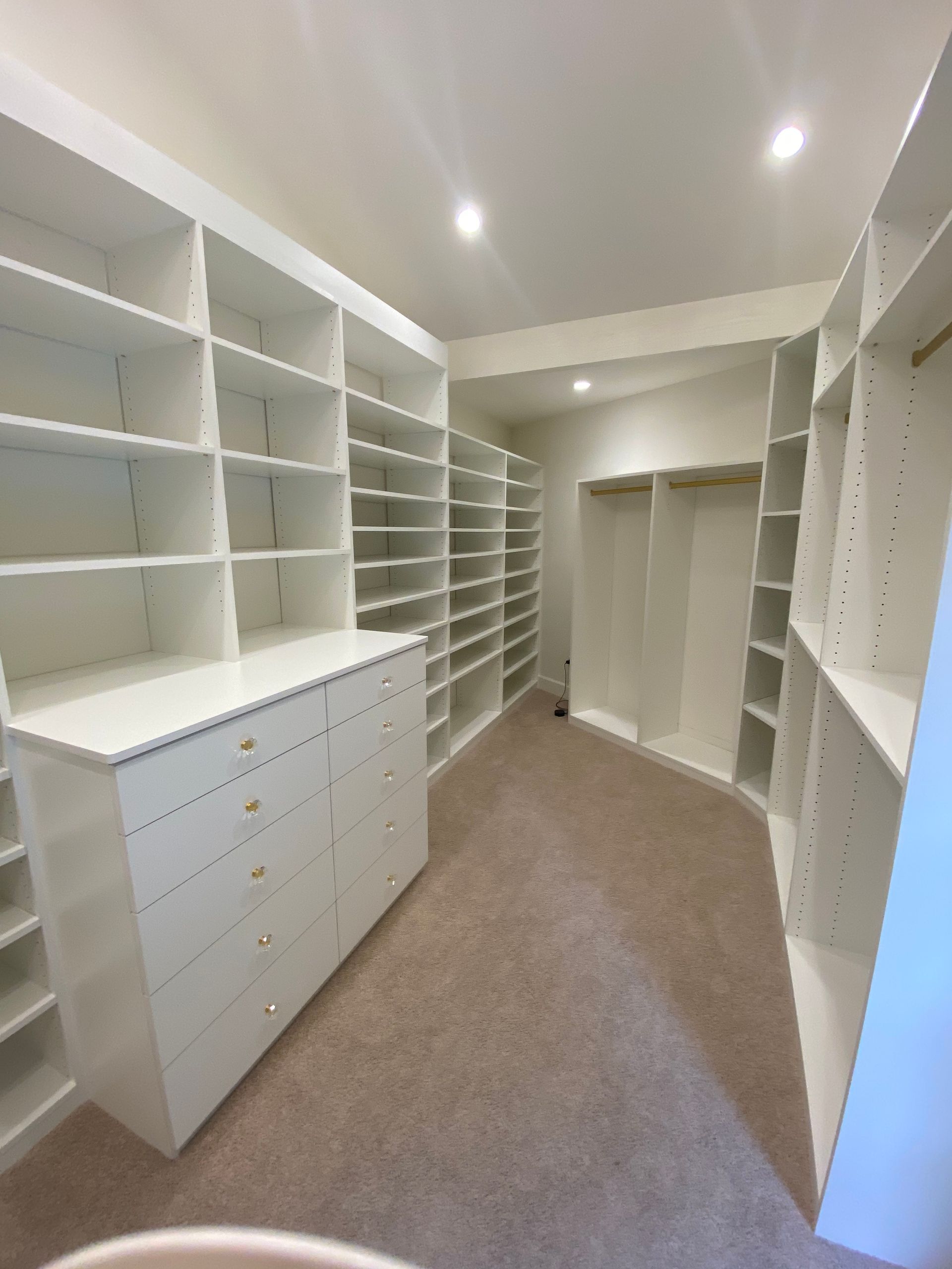 White, built-in closet with shelves, drawers, and hanging rods; beige carpet, bright lighting.