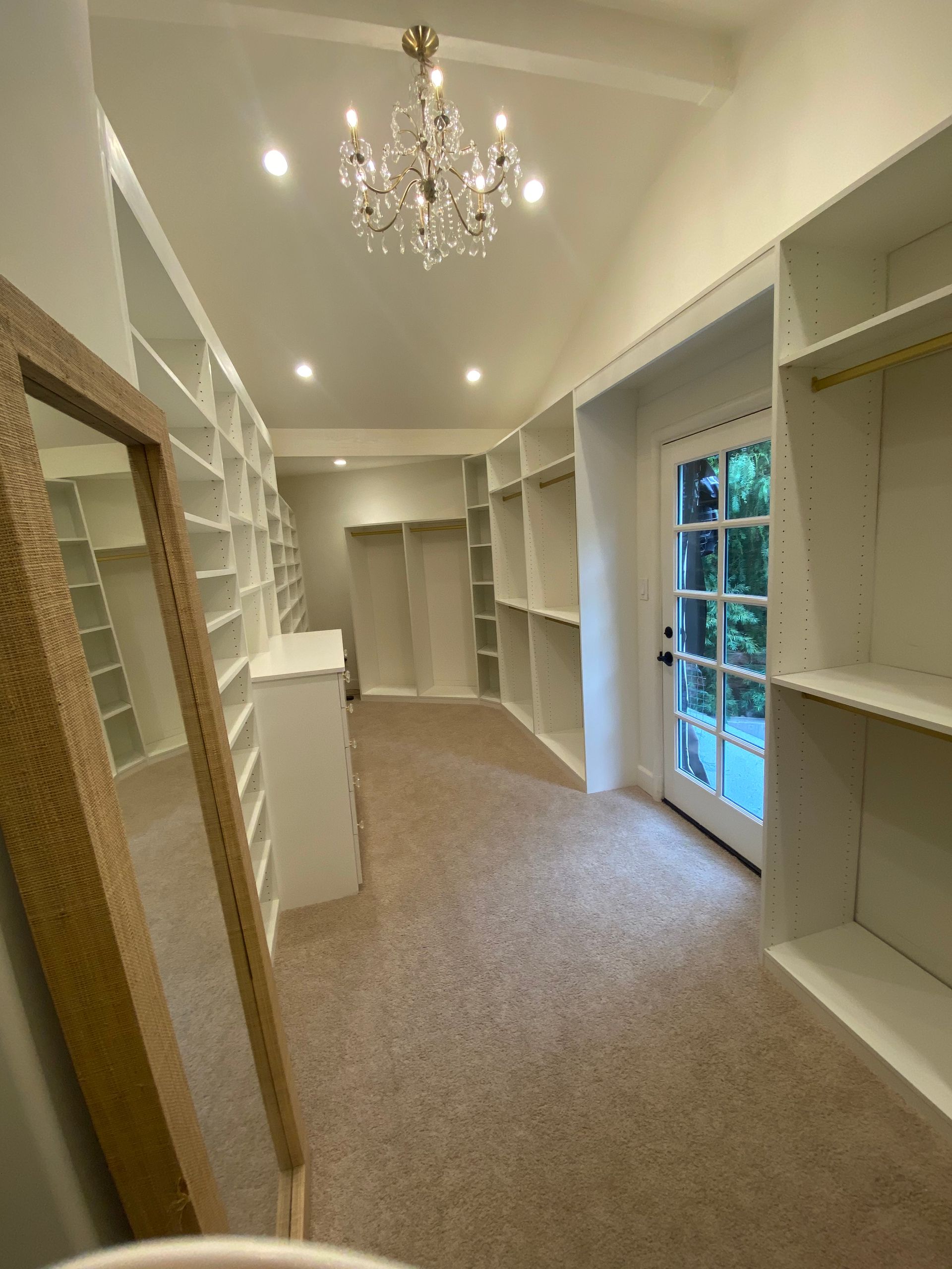 Walk-in closet with white shelves, beige carpet, chandelier, full-length mirror, and door to outside.