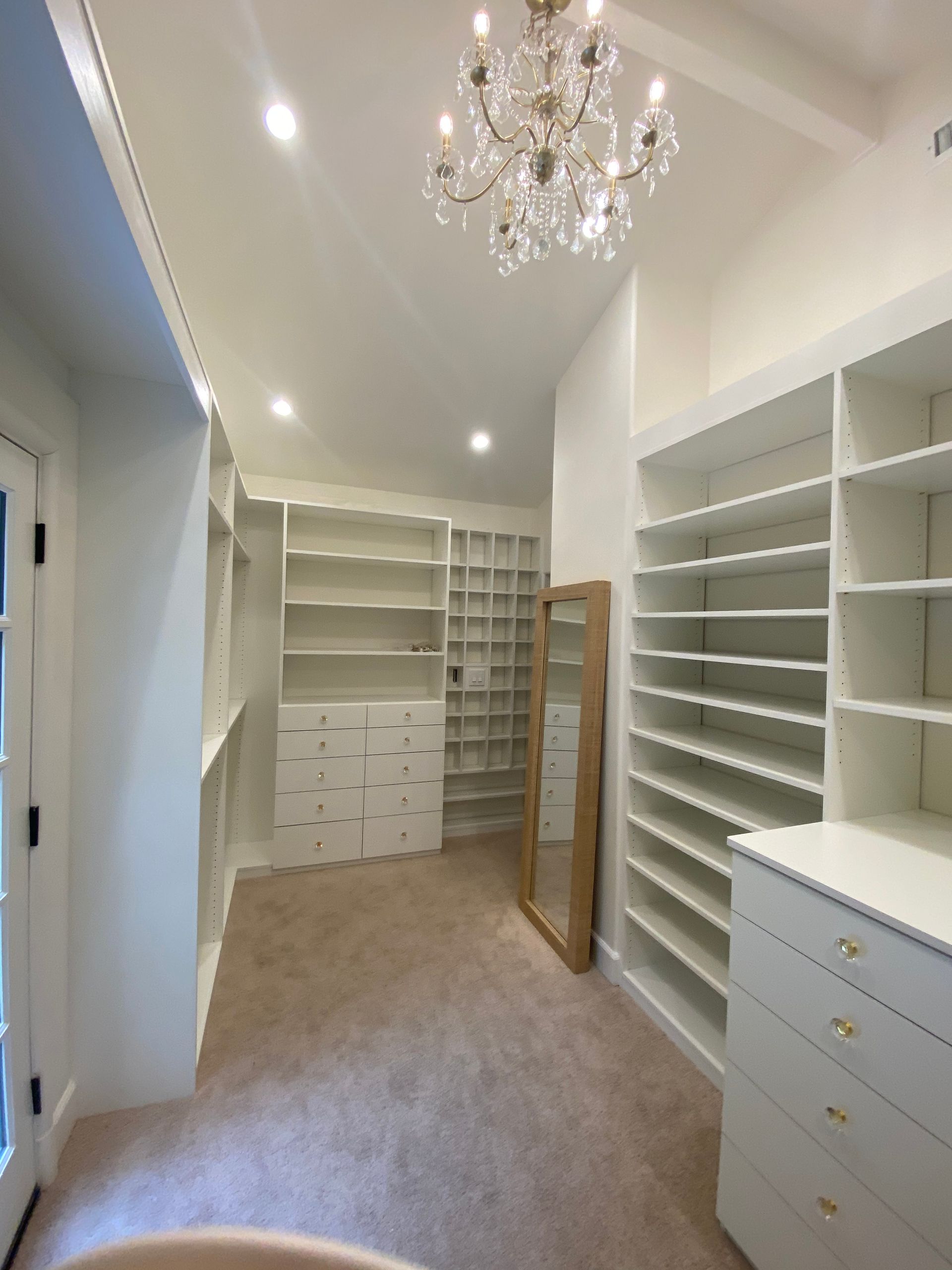 Walk-in closet with white shelves and a chandelier; beige carpet, and a full-length mirror.