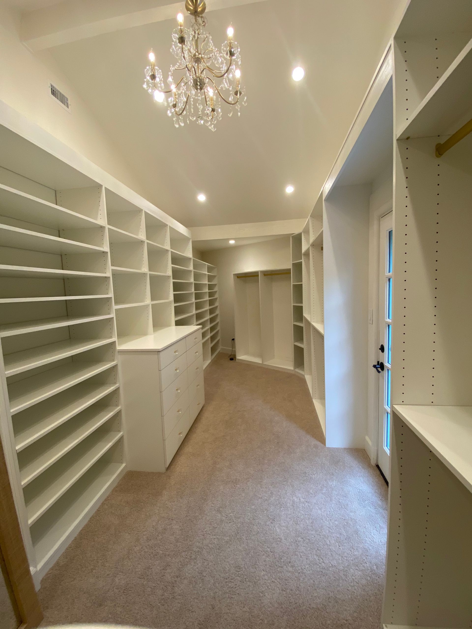 Spacious, bright white walk-in closet with built-in shelves, drawers, and a chandelier; beige carpet.