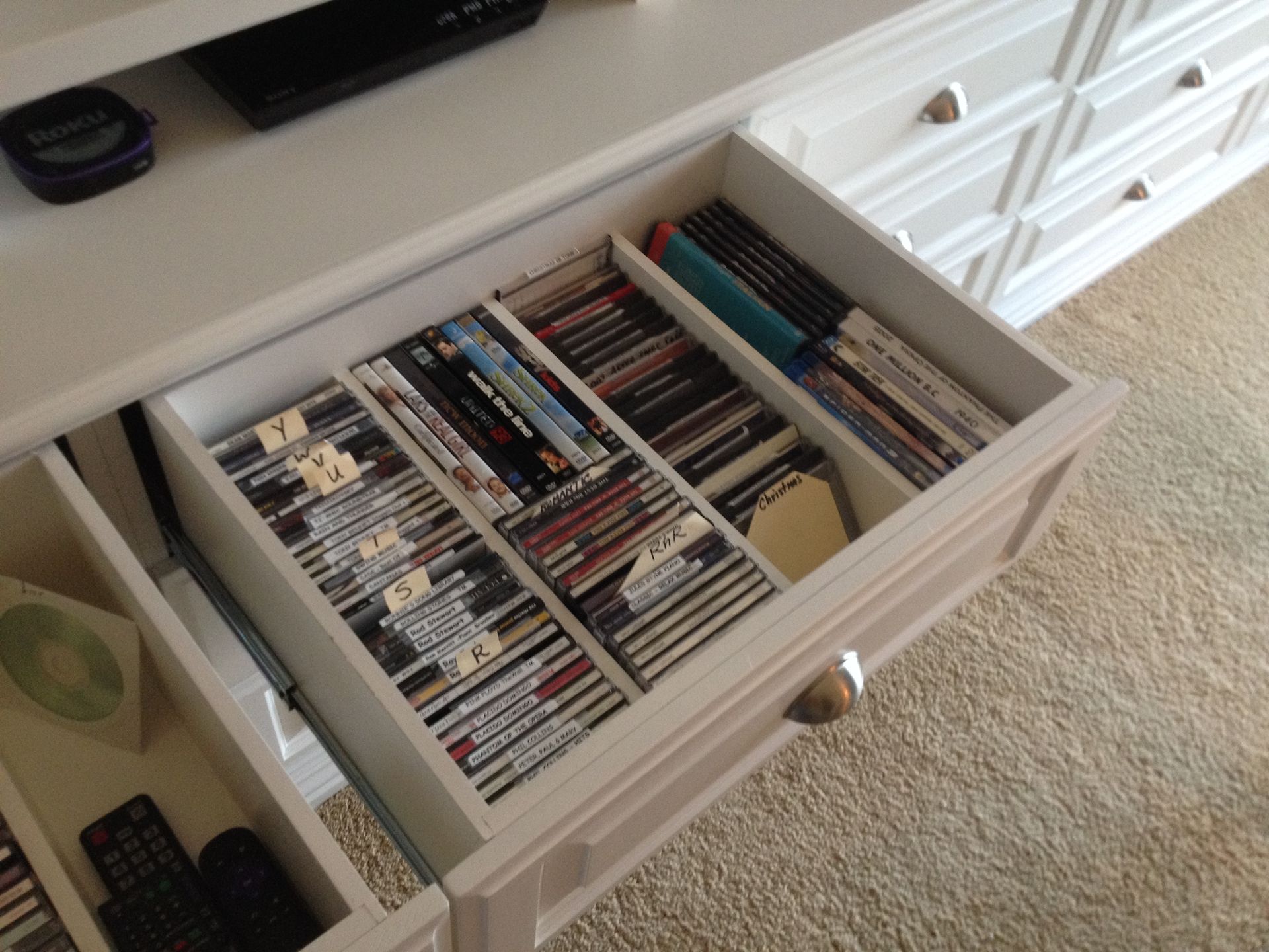 White cabinet drawer open, filled with DVDs, organized with dividers. Another drawer visible with remotes and CDs.