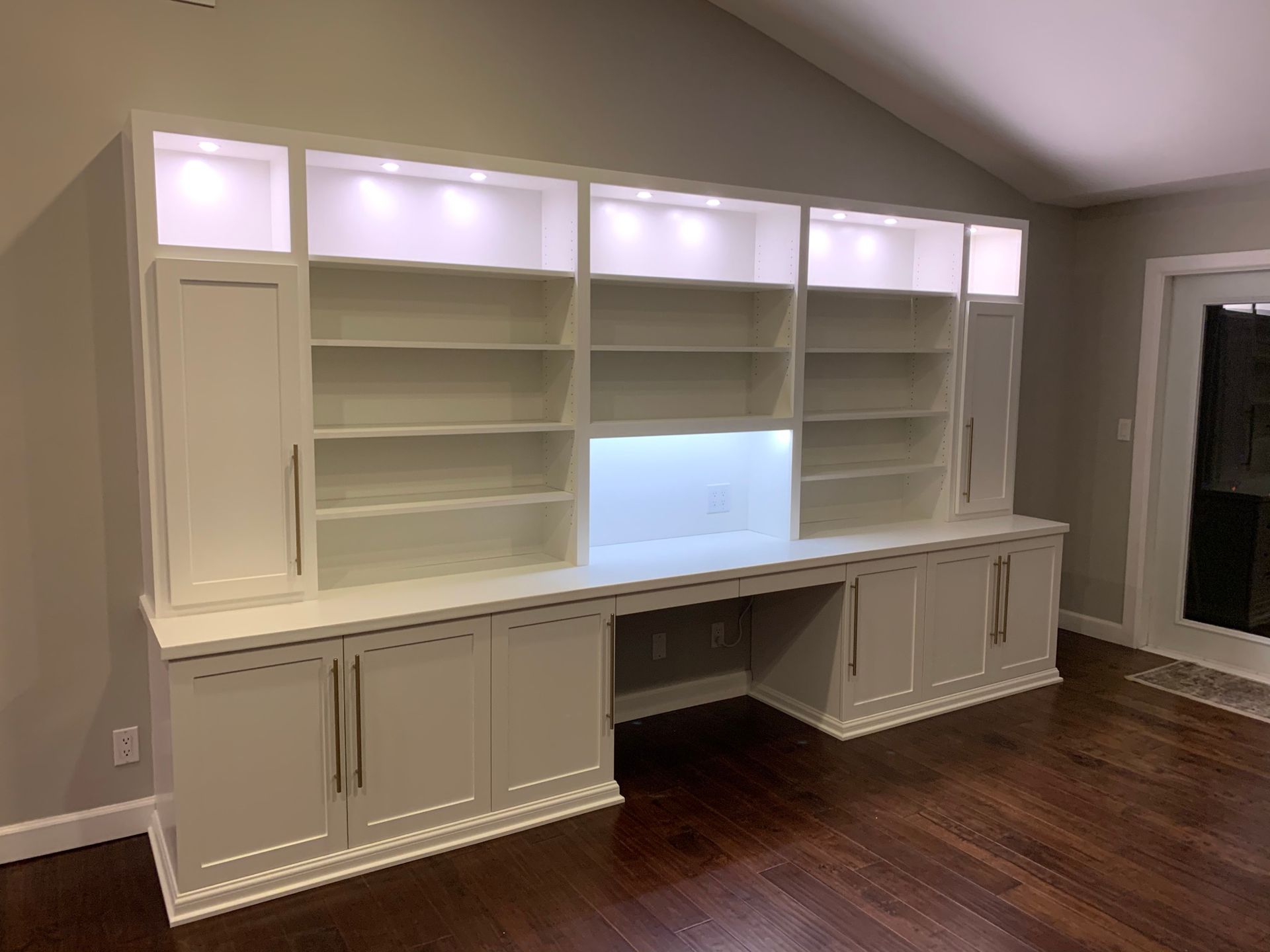 White built-in bookcase with desk, illuminated shelving, and cabinets against a neutral wall; dark wood floor.