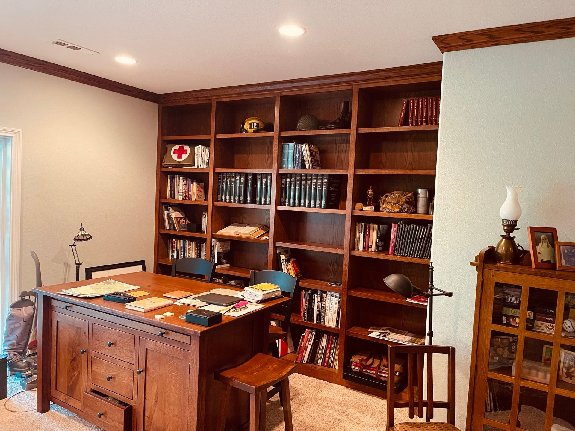 Wooden bookshelves filled with books and various items next to a wooden desk with a lamp, in a room.