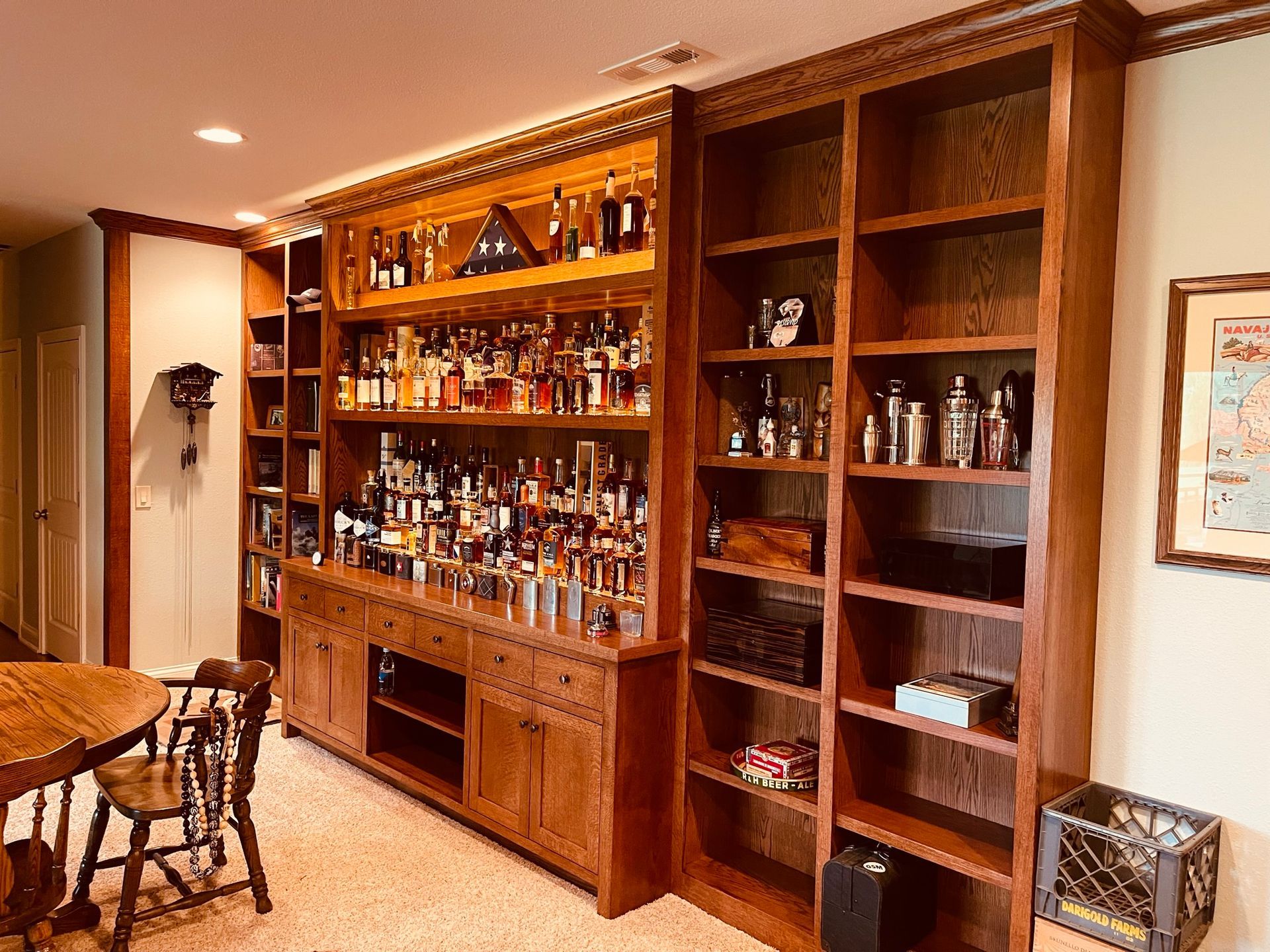 Built-in wooden bar with liquor bottles and shelves, next to a bookcase and a small table.