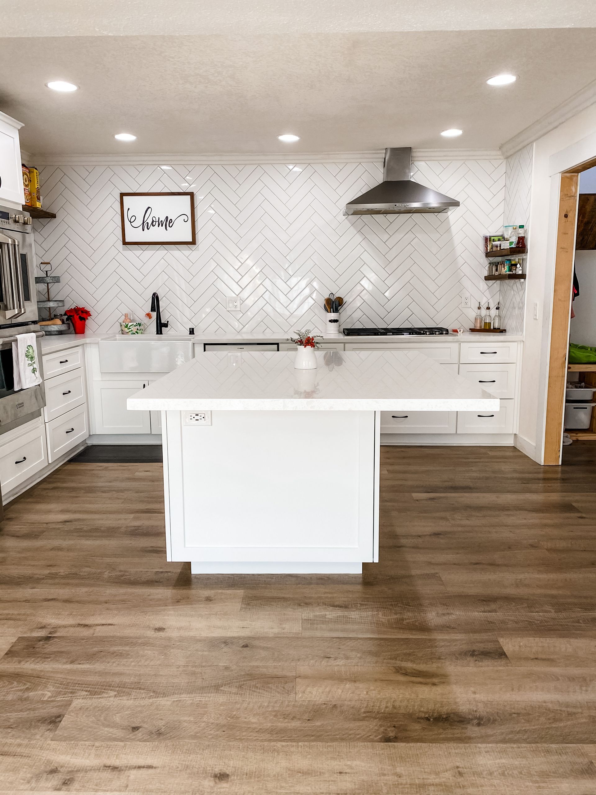 White kitchen with wood floors, island, herringbone backsplash, and stainless steel hood.