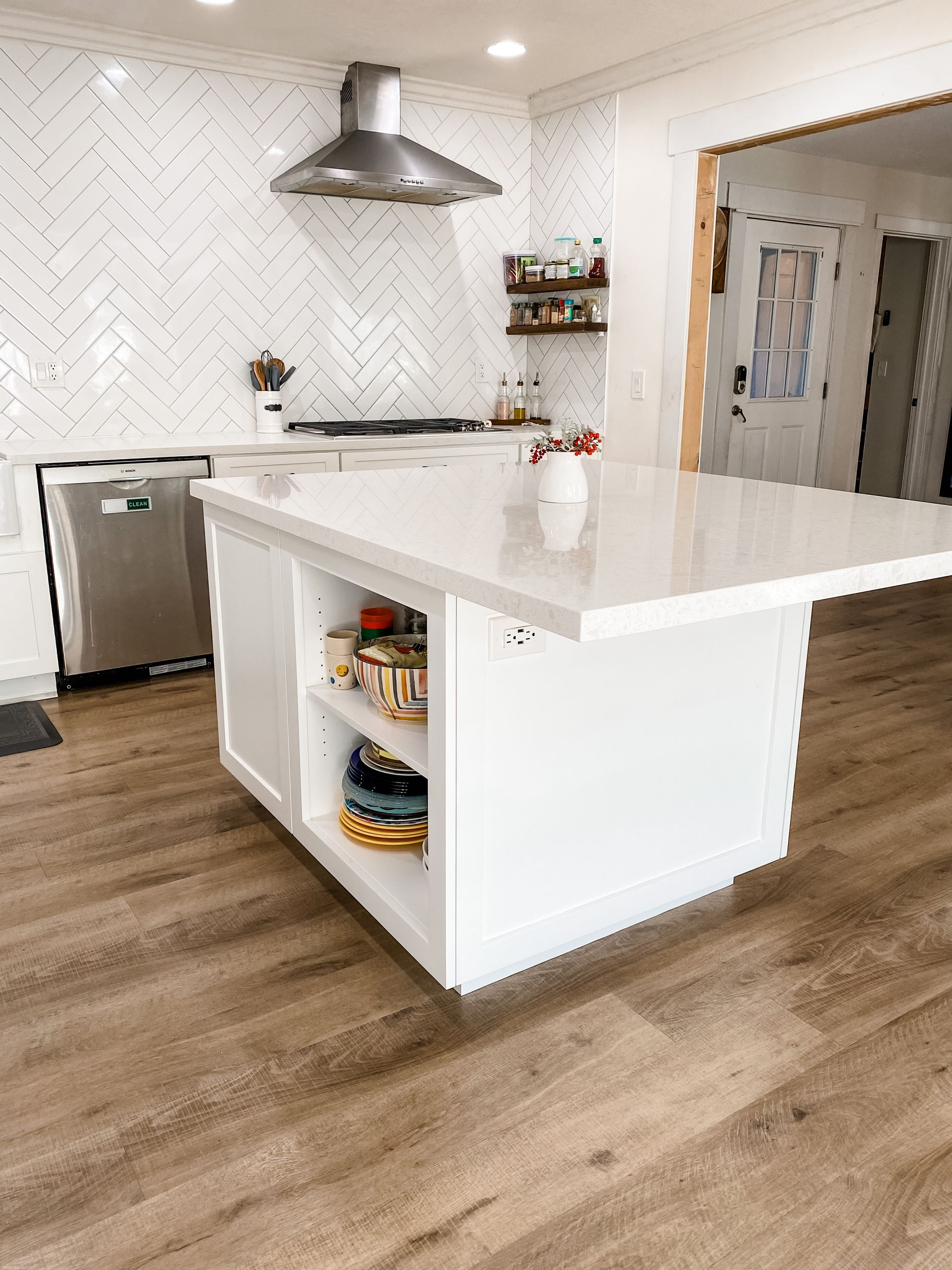 White kitchen with island and herringbone backsplash. Stainless appliances, wood floors, and shelves with decorative items.