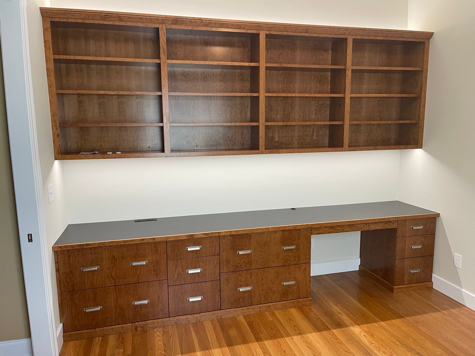 Built-in wooden office desk with drawers and bookshelves. Brown wood, gray desktop. Lit from above.