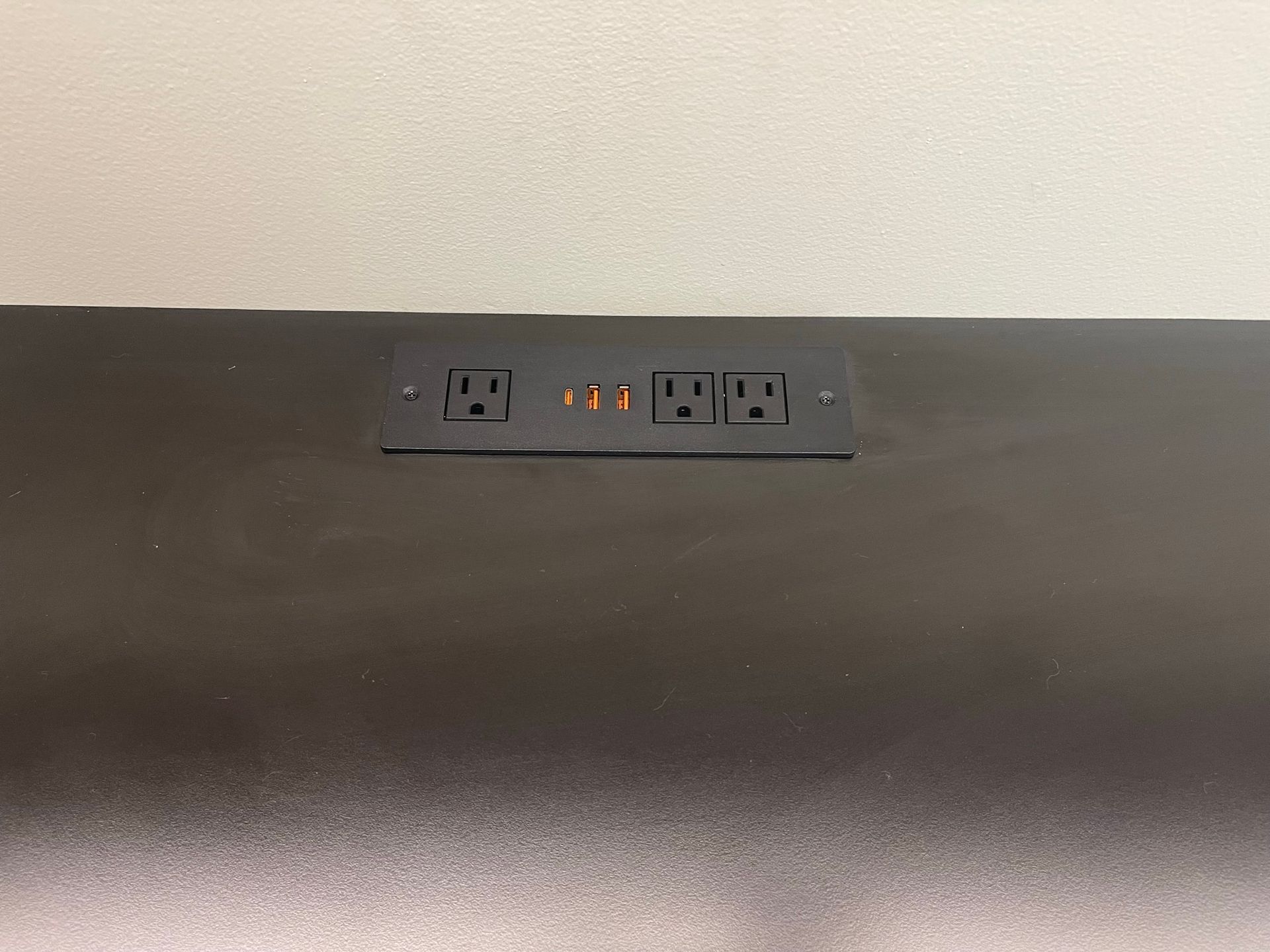 Black desk with electrical outlets and USB ports, flush-mounted, against a white wall.