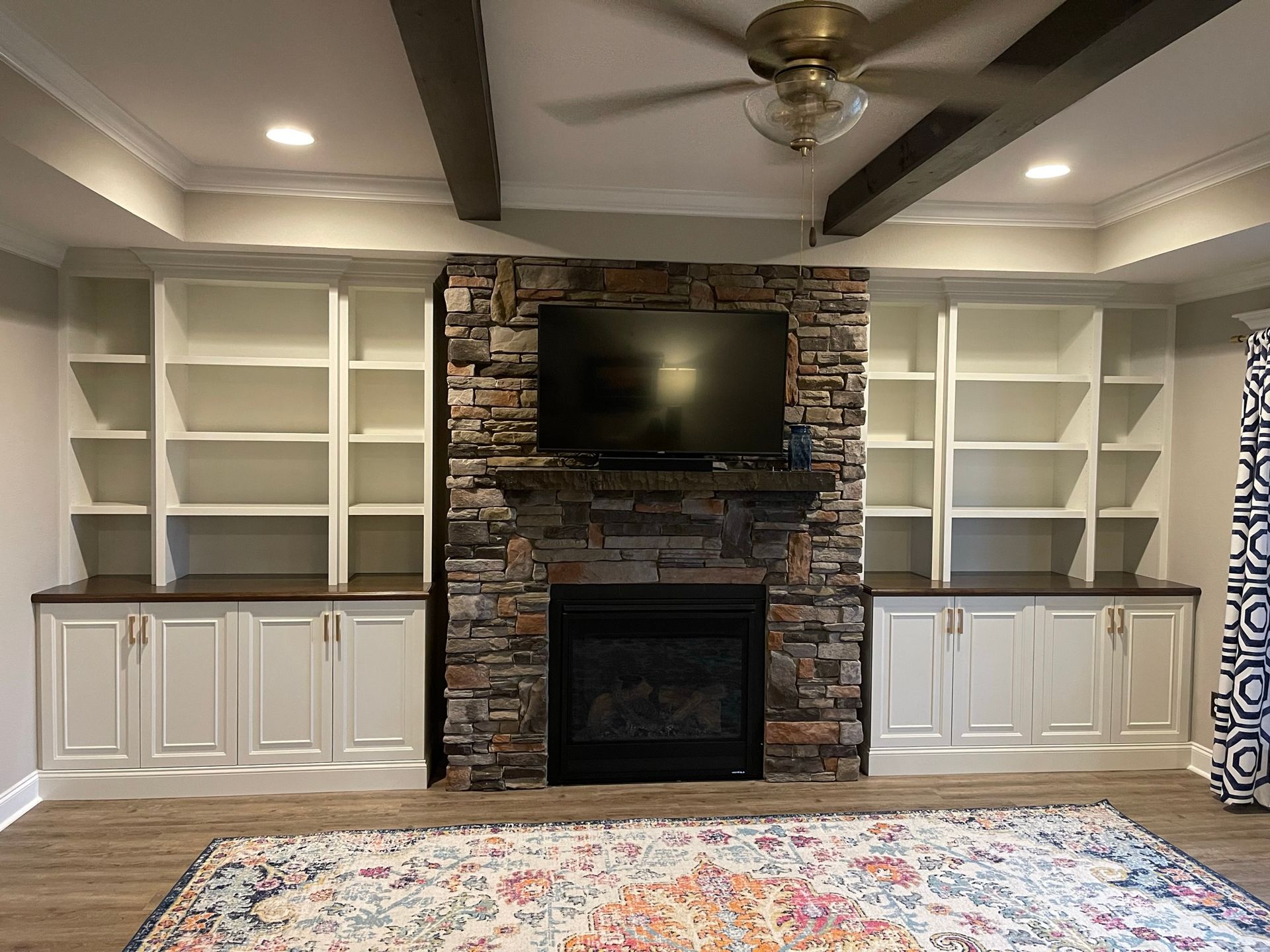 Built-in white bookshelves flank a stone fireplace with a TV above it, in a room with a rug and ceiling beams.