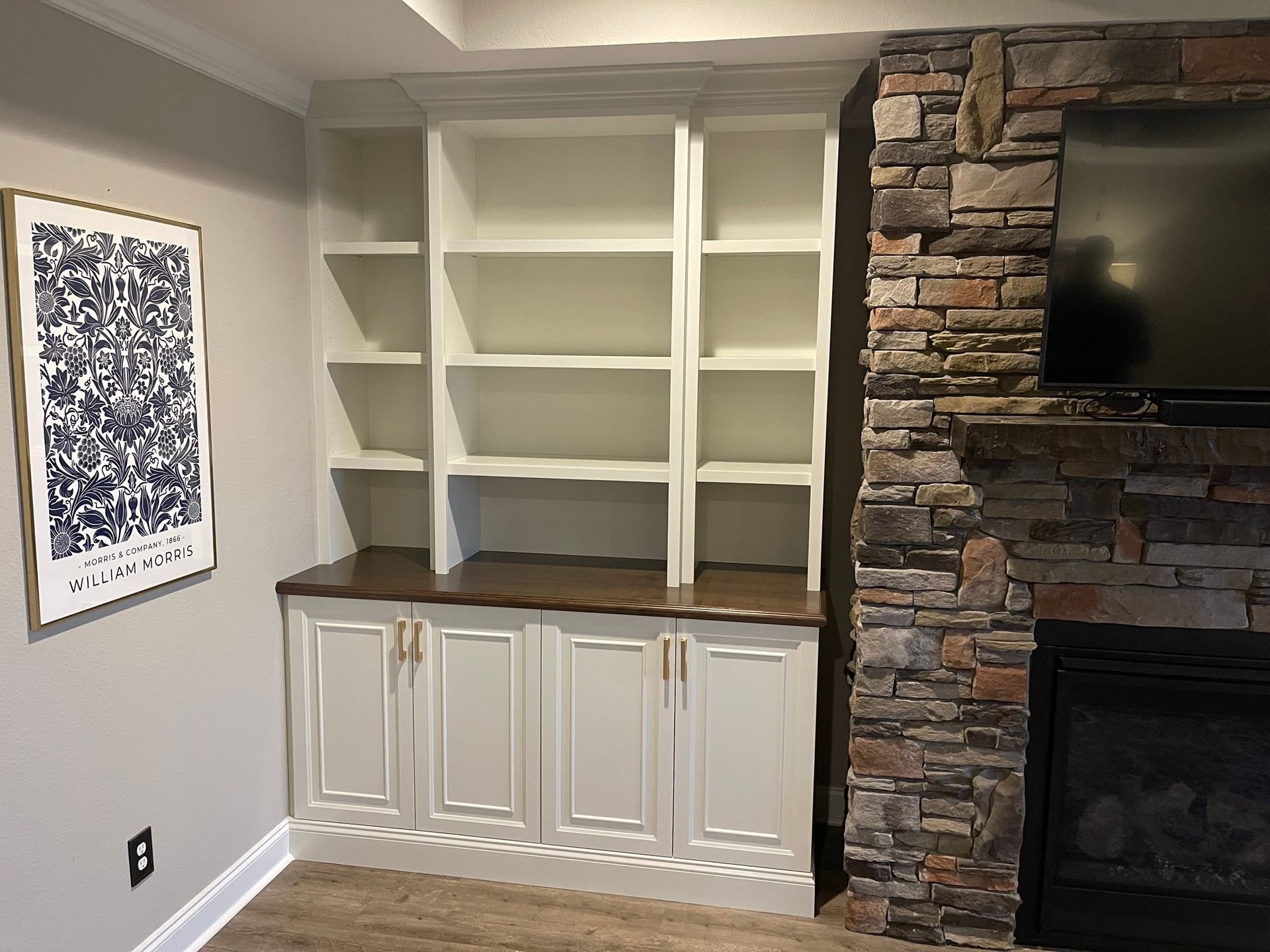 Built-in white bookshelves with cabinets, brown countertop, next to a stone fireplace and artwork.