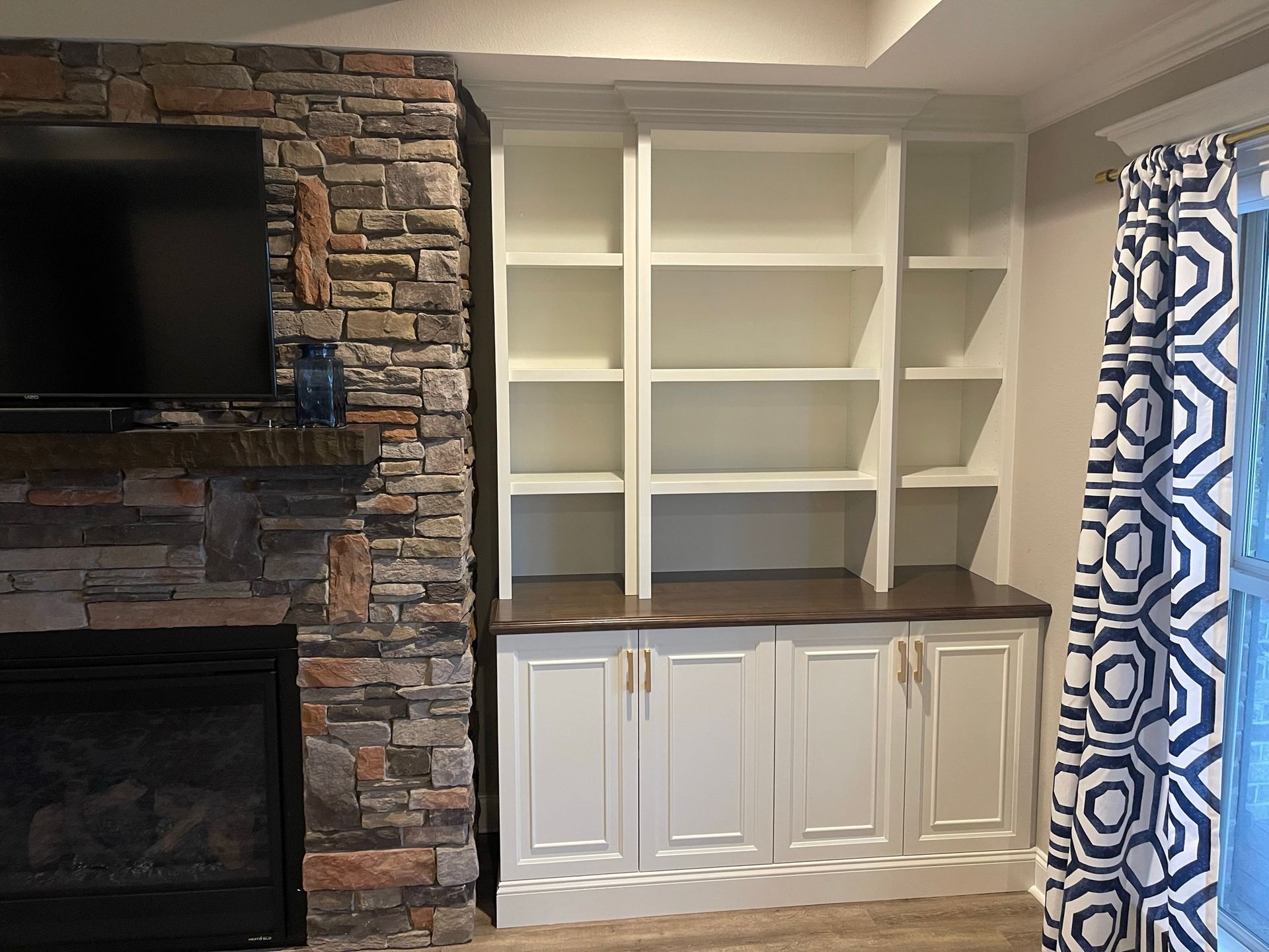 Built-in white bookshelves with cabinets, next to a stone fireplace and patterned curtains.