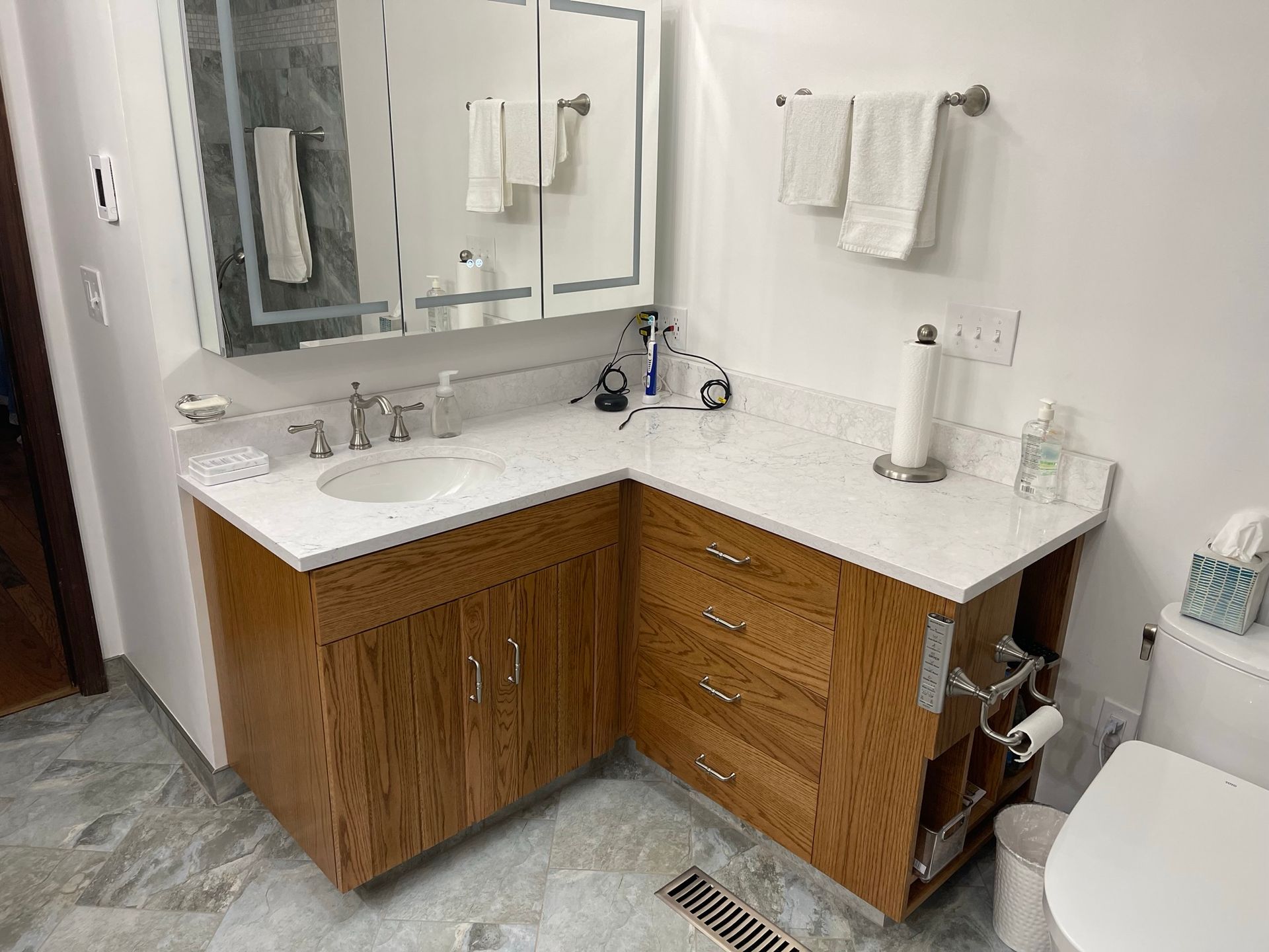 Corner bathroom vanity with white countertop and wooden cabinets, mirrored medicine cabinet, and toilet.