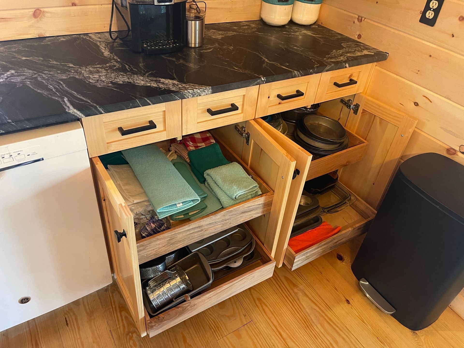 Kitchen cabinets with open drawers. One drawer holds towels, another has pans. A black countertop holds a coffee maker.