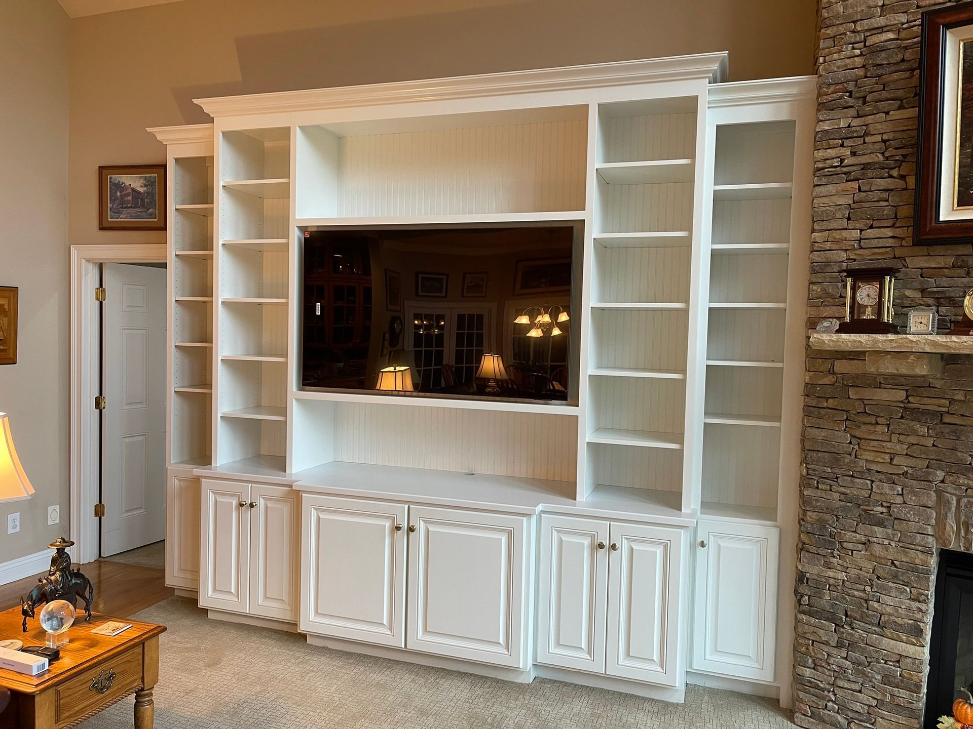 White built-in entertainment center with a TV, cabinets, and shelves. Adjacent to a stone fireplace.