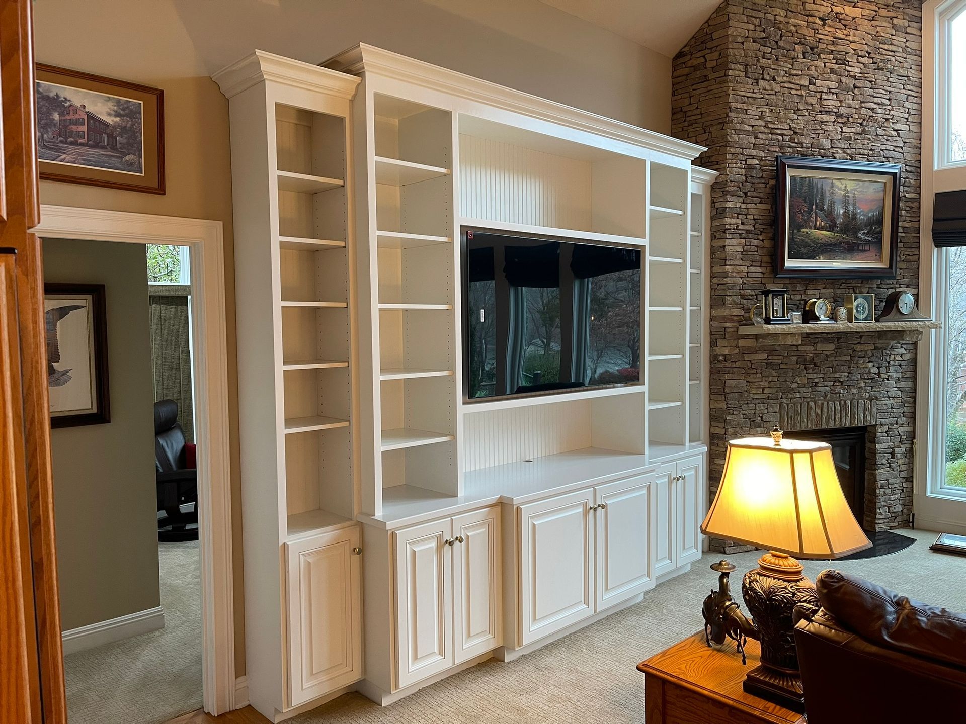Built-in white entertainment center with cabinets, shelves, and a TV; fireplace and painting in the background.