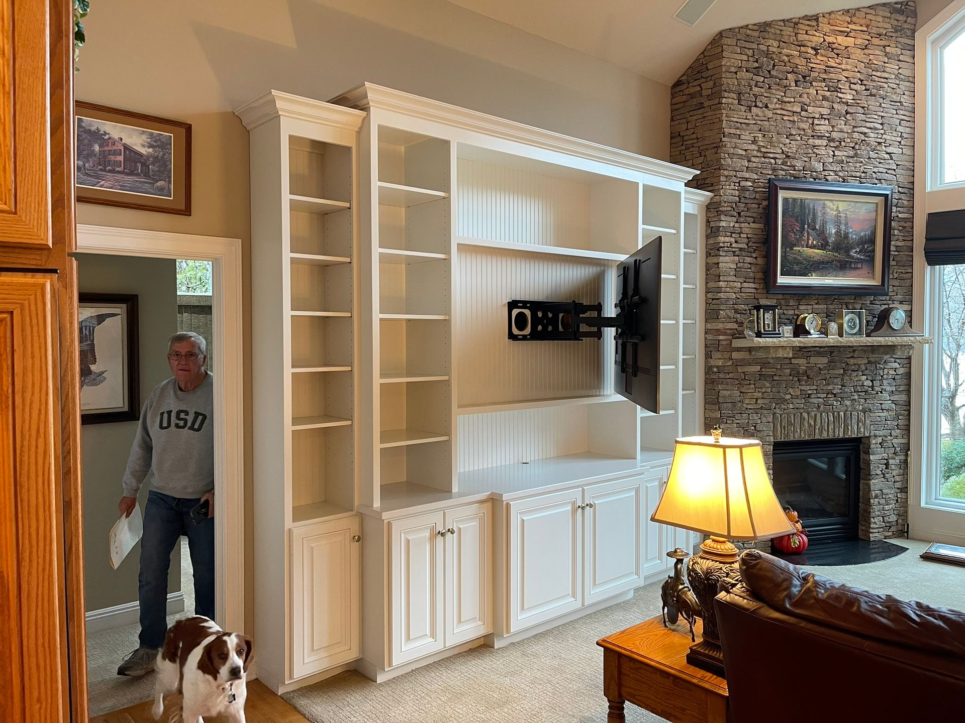 Built-in white shelves with TV mount; man and dog in doorway, fireplace in background.