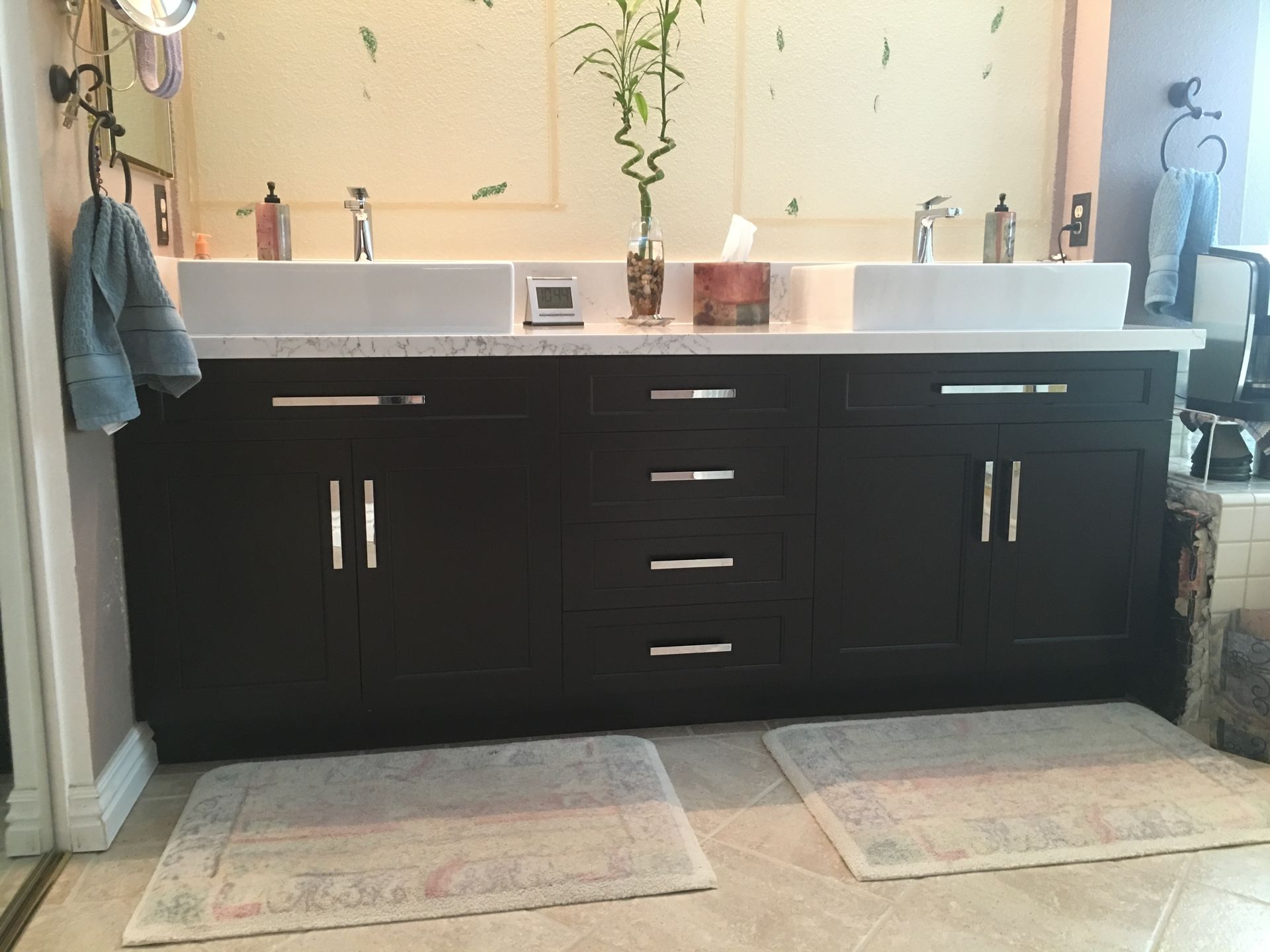 Dark brown bathroom vanity with white countertop and sinks, with bamboo in the center. Two rugs on the floor.