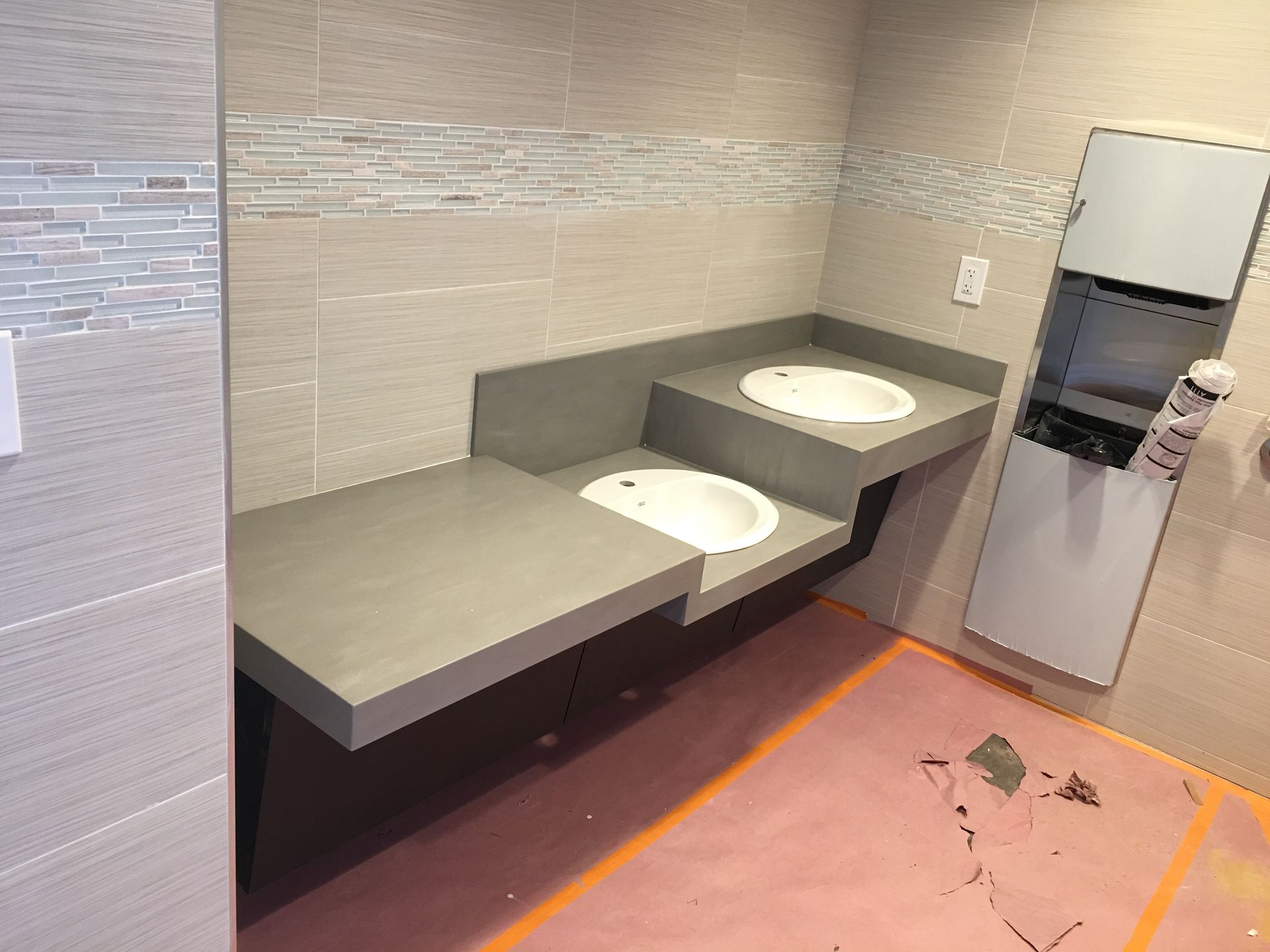 Bathroom with two sinks on stepped counter, tiled walls, and a paper towel dispenser.