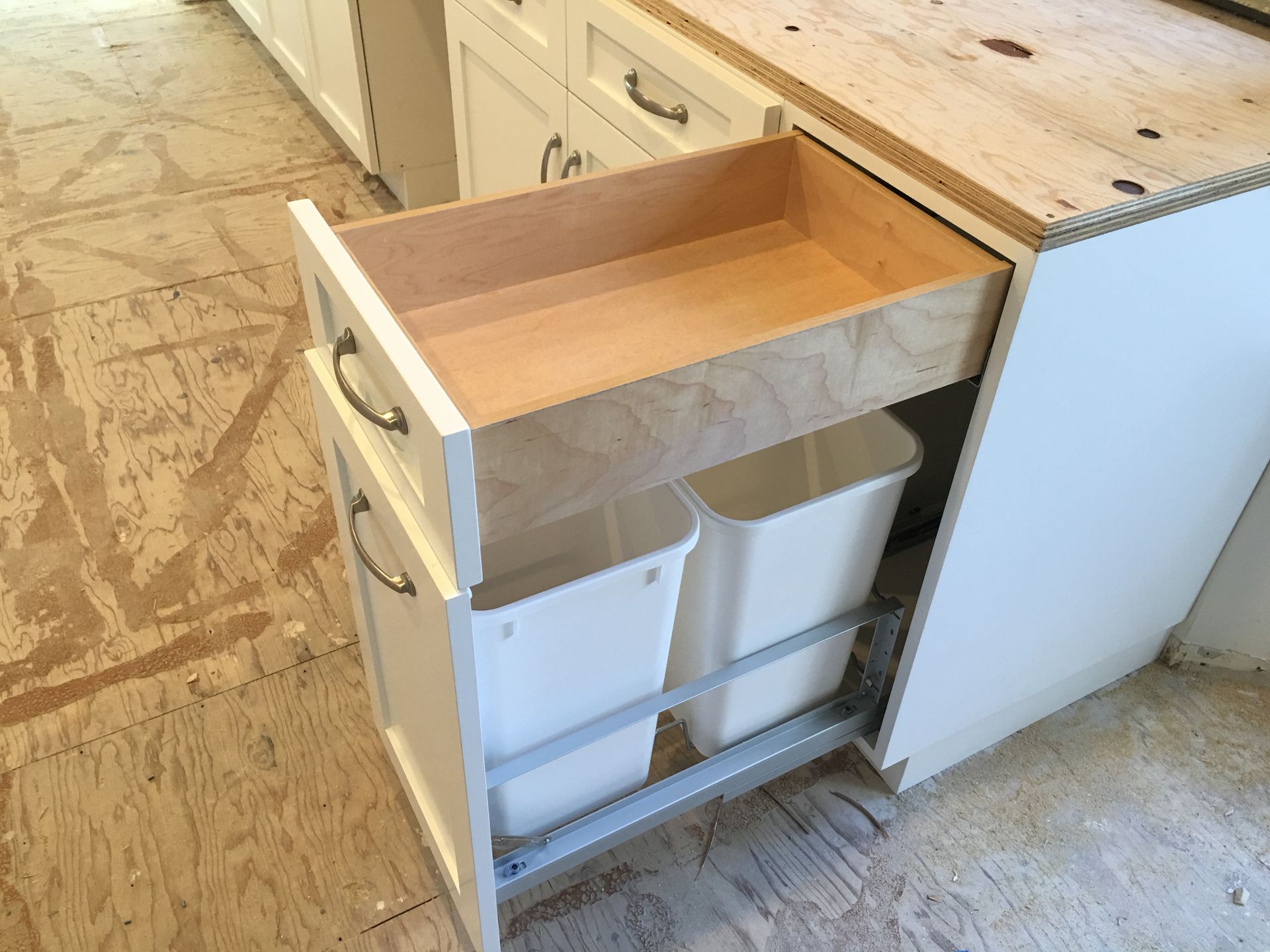 Kitchen cabinet with pull-out trash bins. White cabinet, light wood drawer. Two white trash bins.
