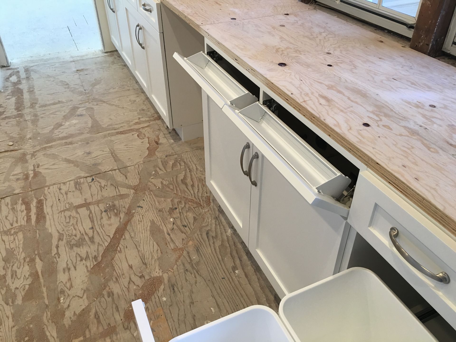 White kitchen cabinets with pull-out trash bin under a wooden countertop. Interior under construction.