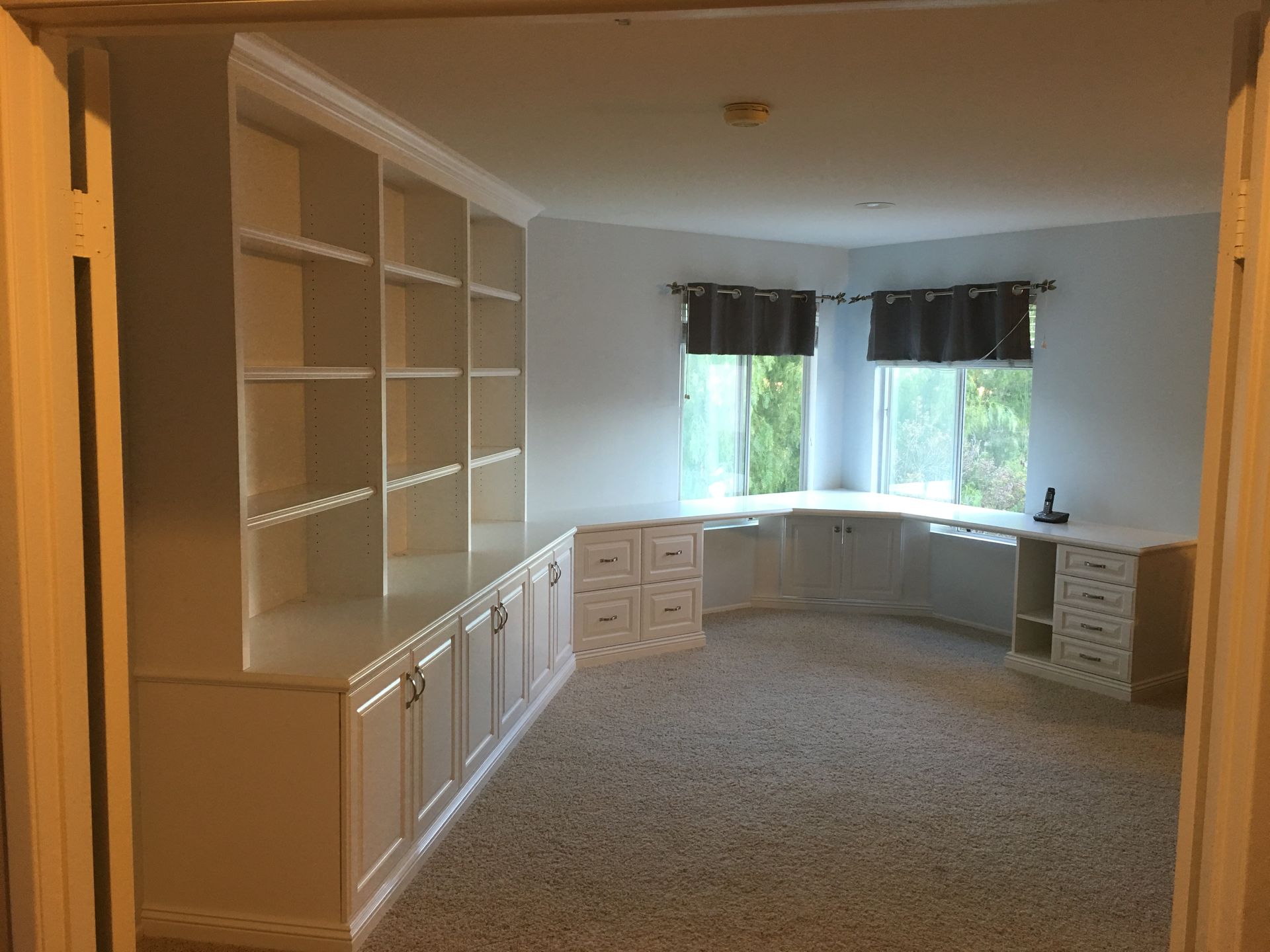 Built-in white bookshelves and cabinets in a room with a corner desk and windows, light carpet.