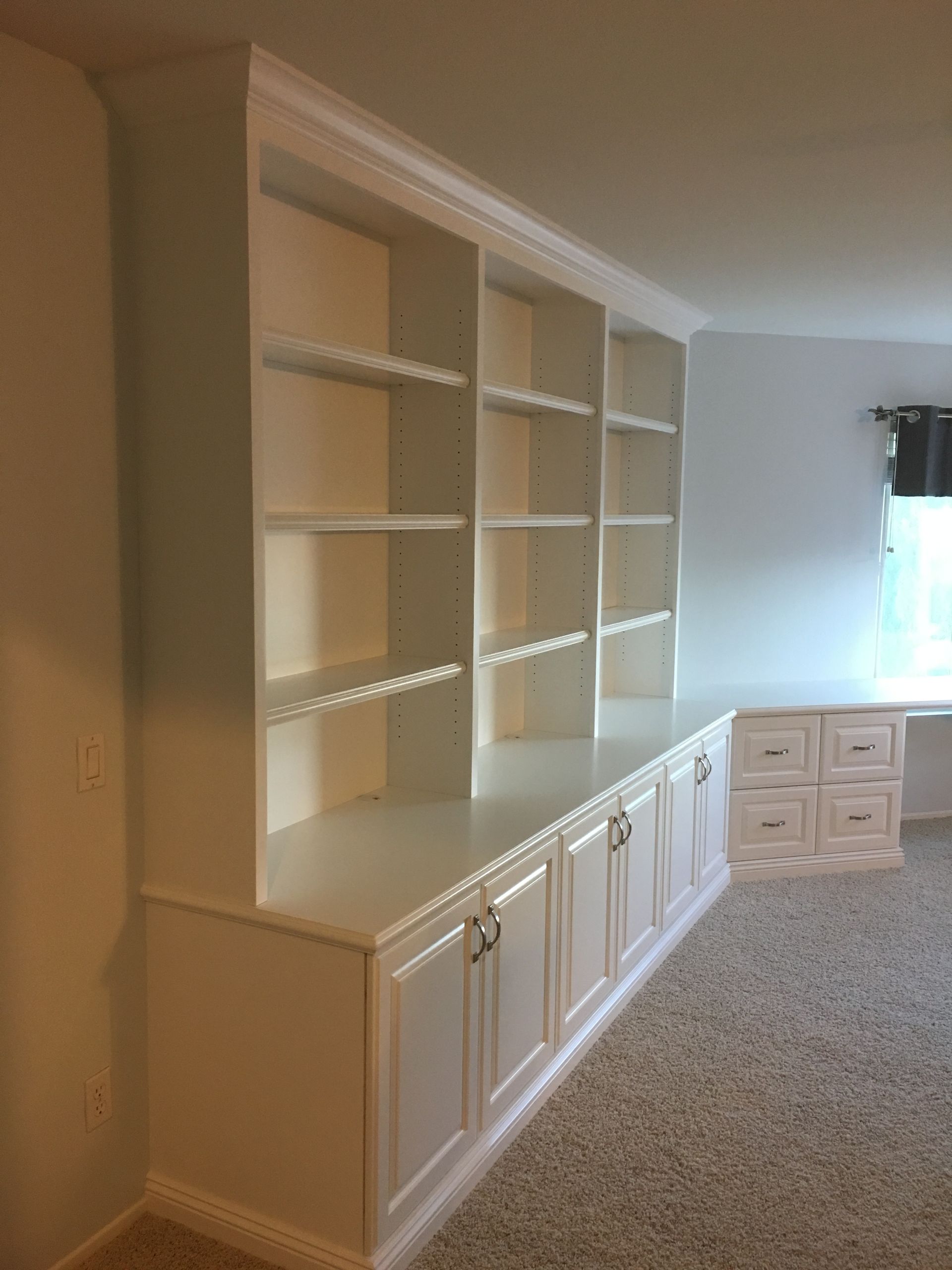 White built-in bookcase with shelves and cabinets, against a white wall and carpeted floor.