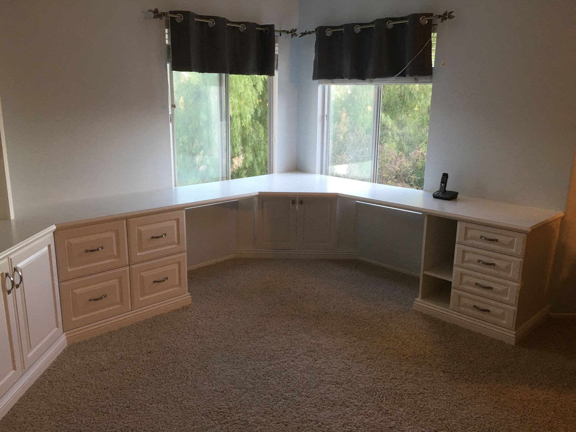 Built-in white desk with drawers and cabinets in a room with a bay window and dark curtains.