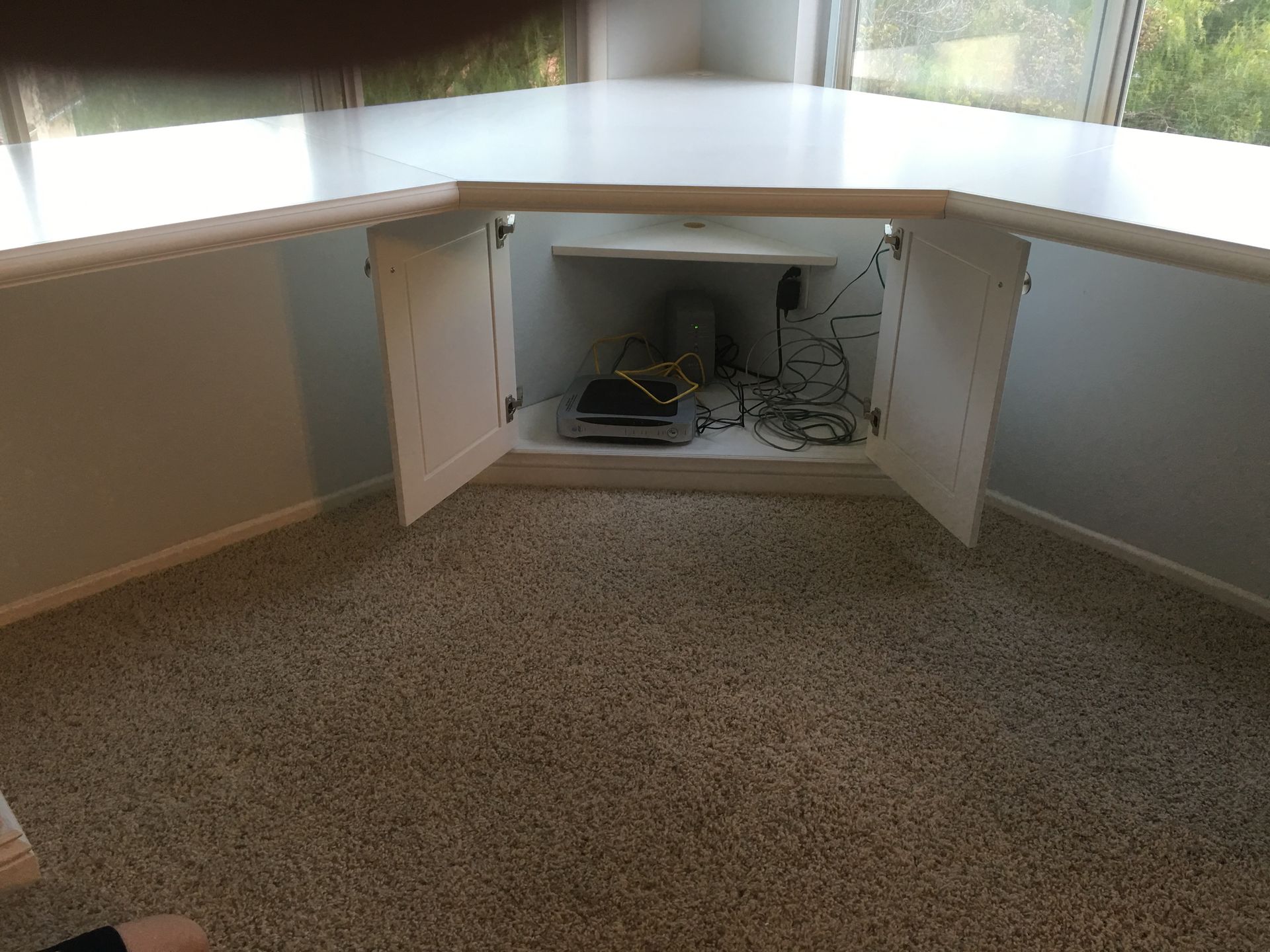 Built-in white desk with cabinets, housing a computer and cables, in a corner of a room with carpet.